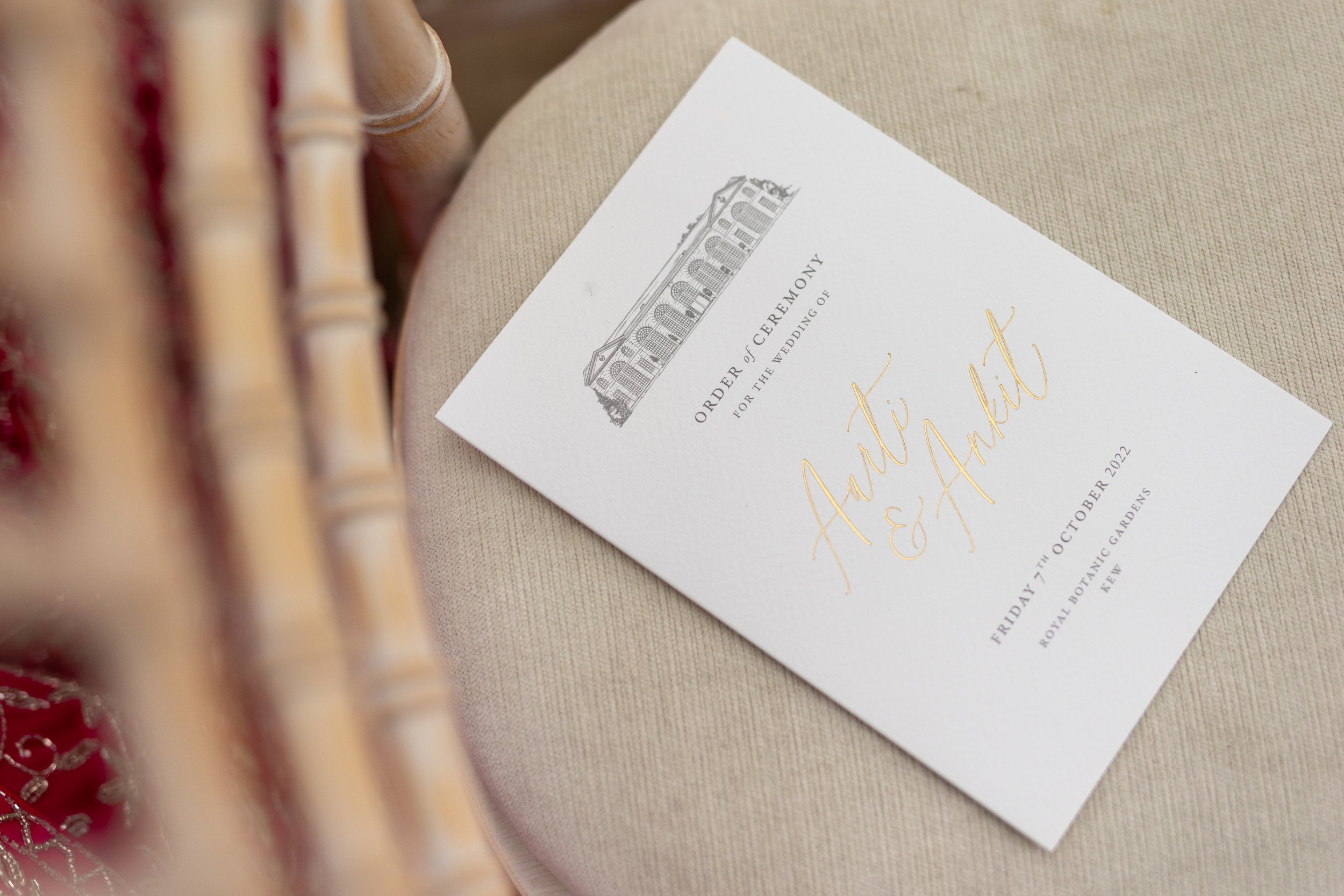 Wedding order of ceremony invitation with an illustration of a building at the top, gold script with the names Arti and Amani, and event details including date, location, and venue on a beige fabric surface.