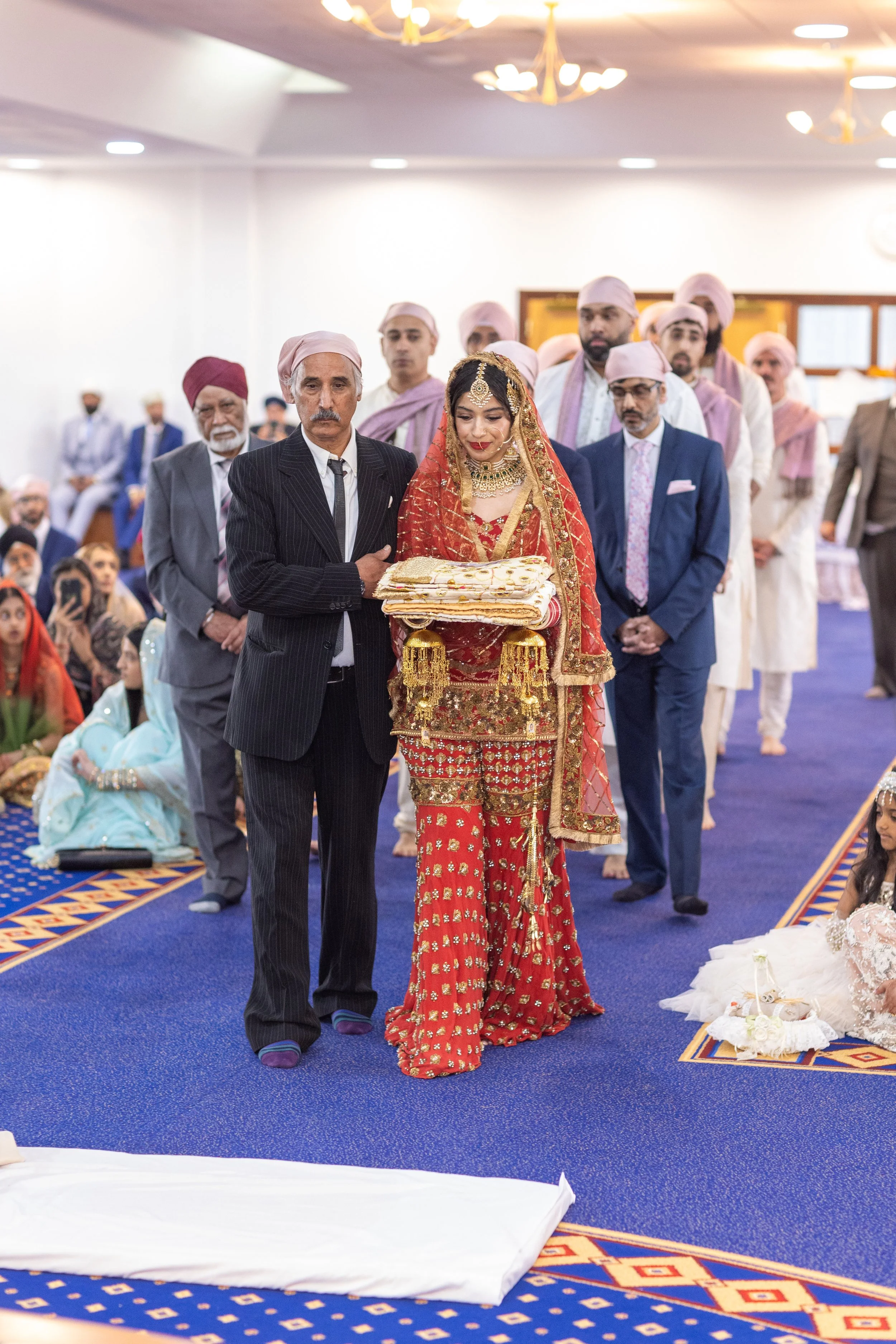 A bride dressed in traditional Indian wedding attire, holding a folded cloth, walking with an older man, surrounded by family and friends in a wedding ceremony.
