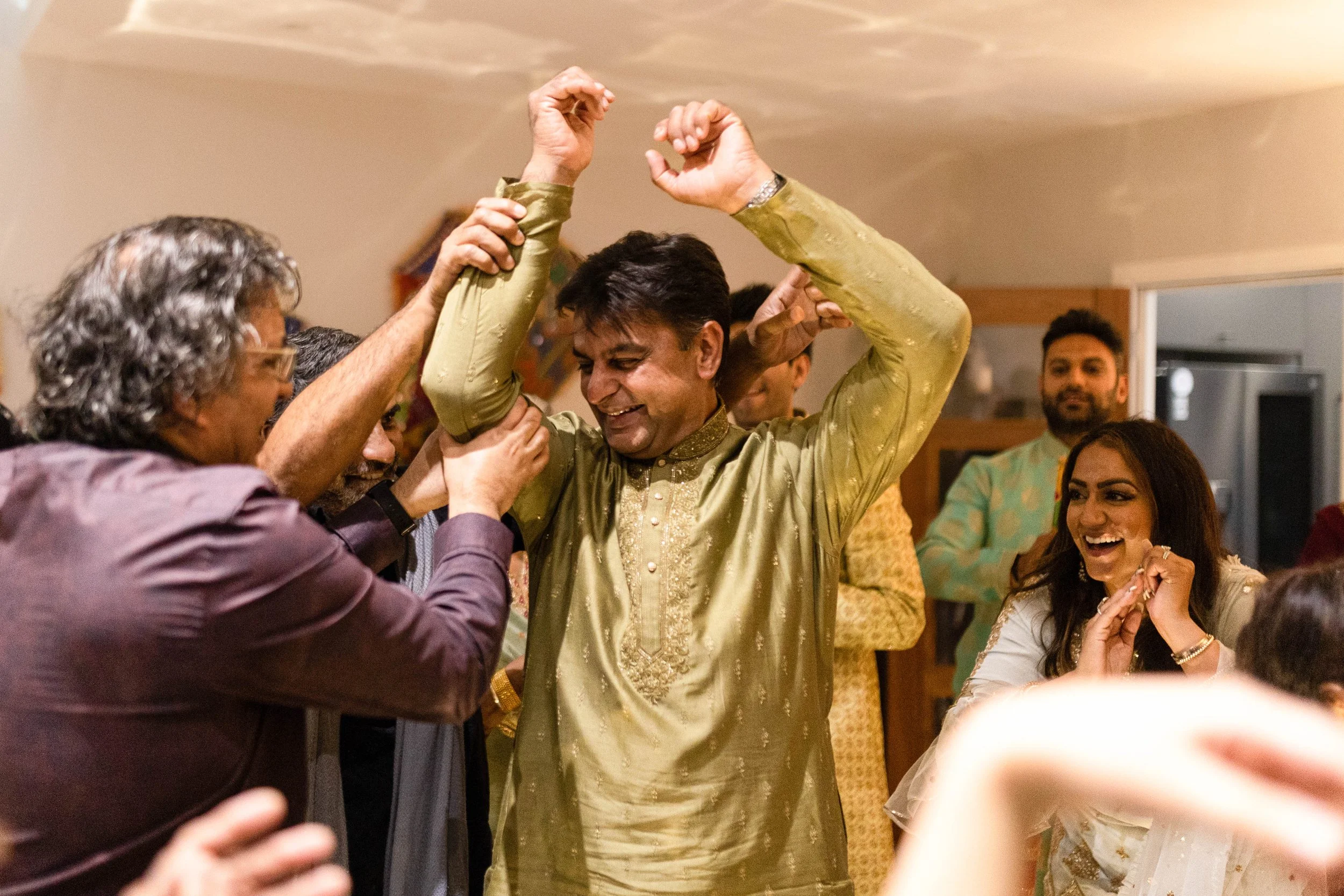 People celebrating at a gathering, with a man in traditional Indian attire smiling as others around him cheer and raise their hands.