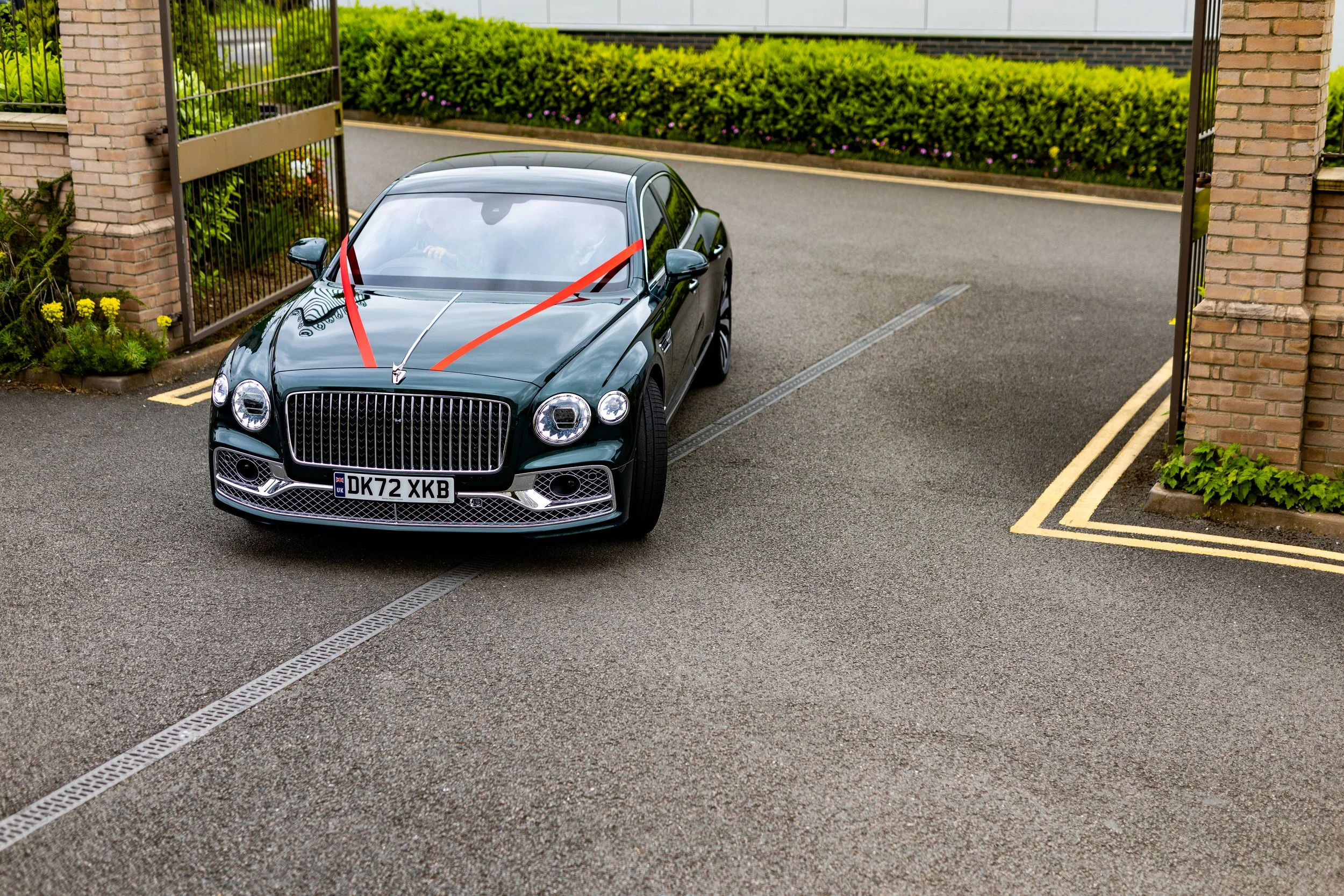 A black luxury sedan with a red ribbon on the hood, parked at the entrance of a gated residential area.