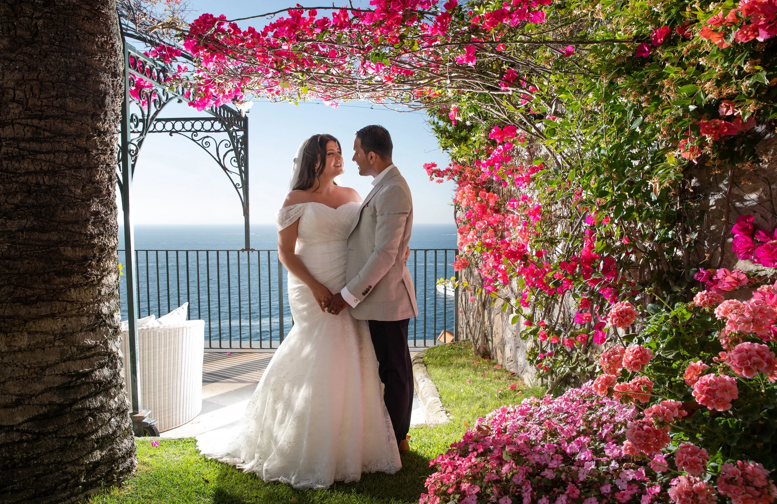 A bride and groom holding hands and looking at each other on a balcony overlooking the ocean, surrounded by pink and green flowering plants and a tree trunk.