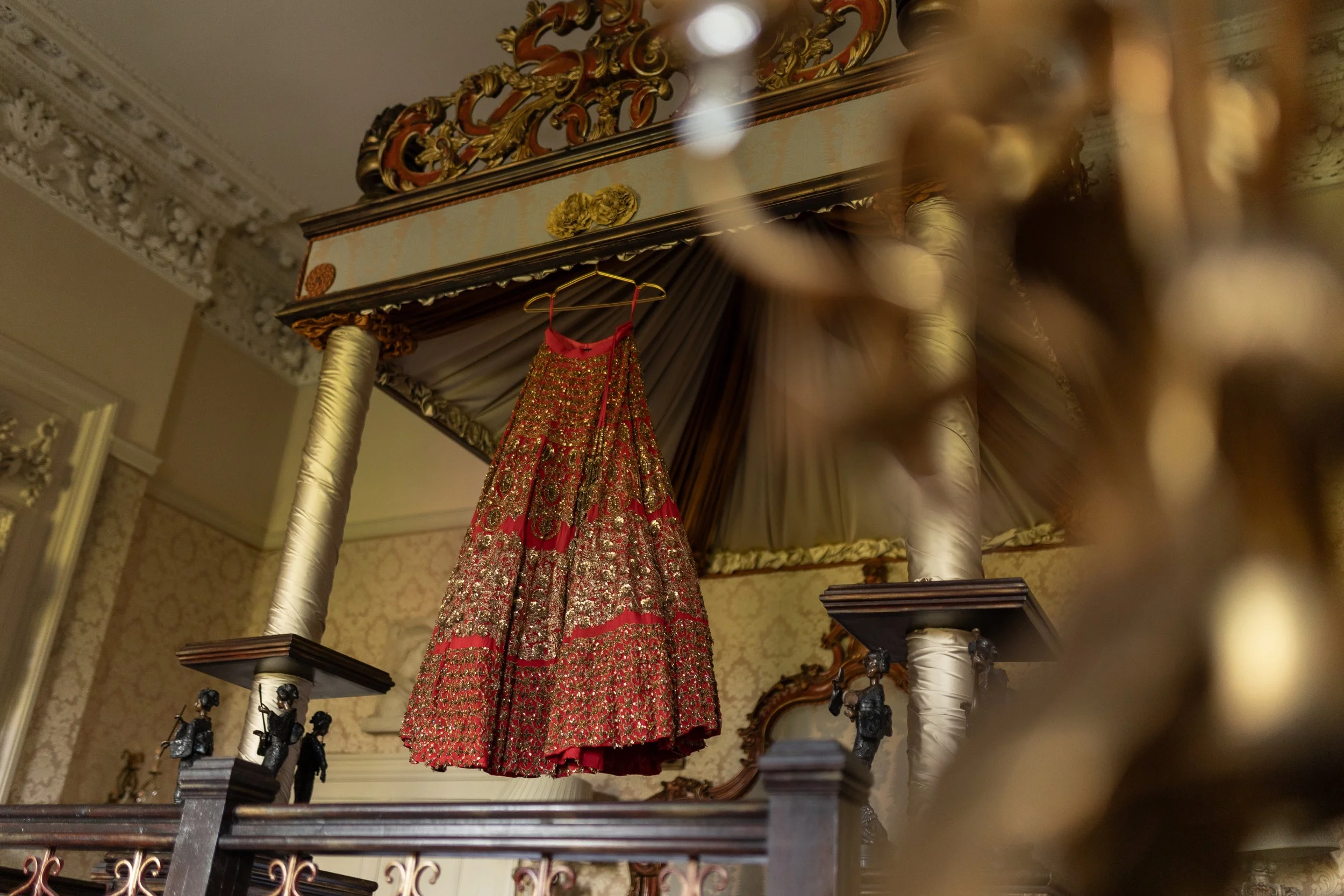A red and gold ornate dress hanging from a hook inside a lavishly decorated room with intricate crown molding and wallpaper, displayed beneath a decorative canopy with gold and brown accents.