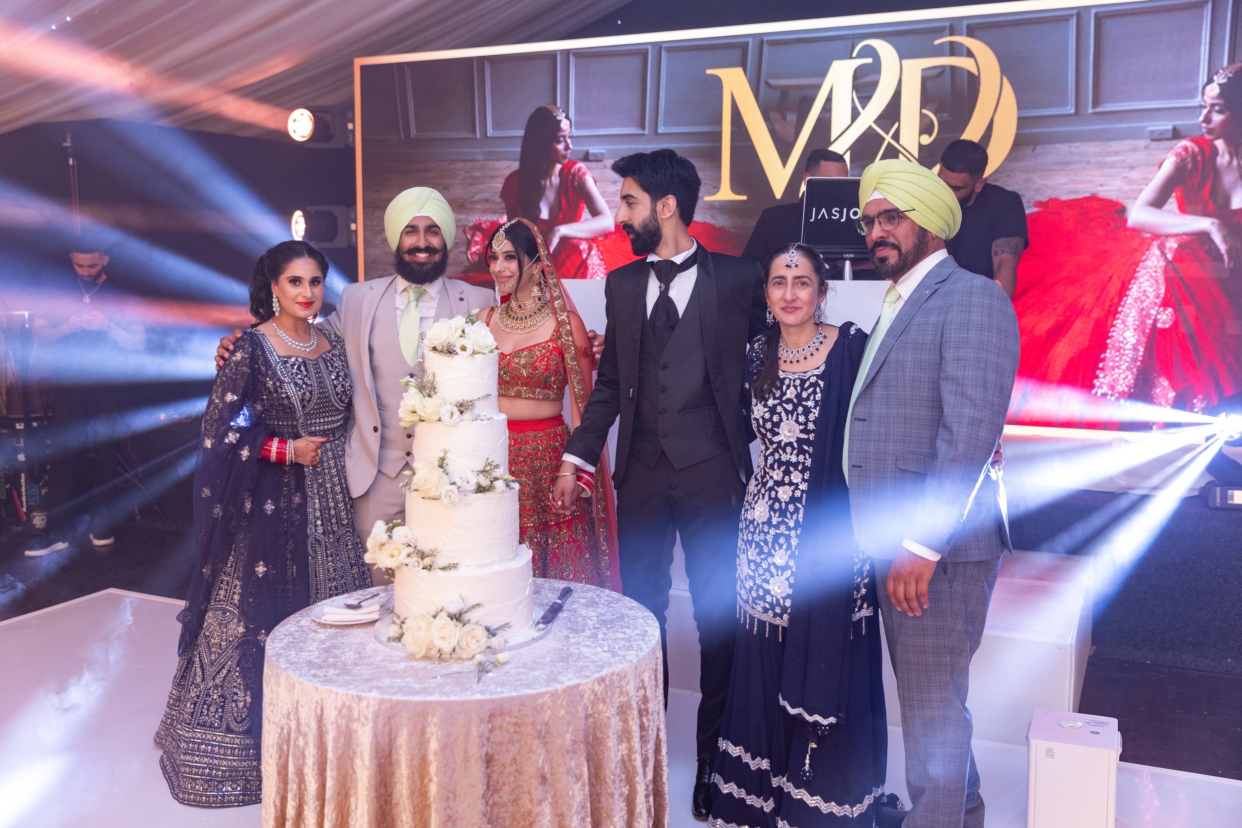 Group of people celebrating a wedding with a multi-tiered white wedding cake decorated with white roses, all dressed in traditional Indian attire and formal wear, standing in front of a stage with lights and a large screen displaying the wedding init
