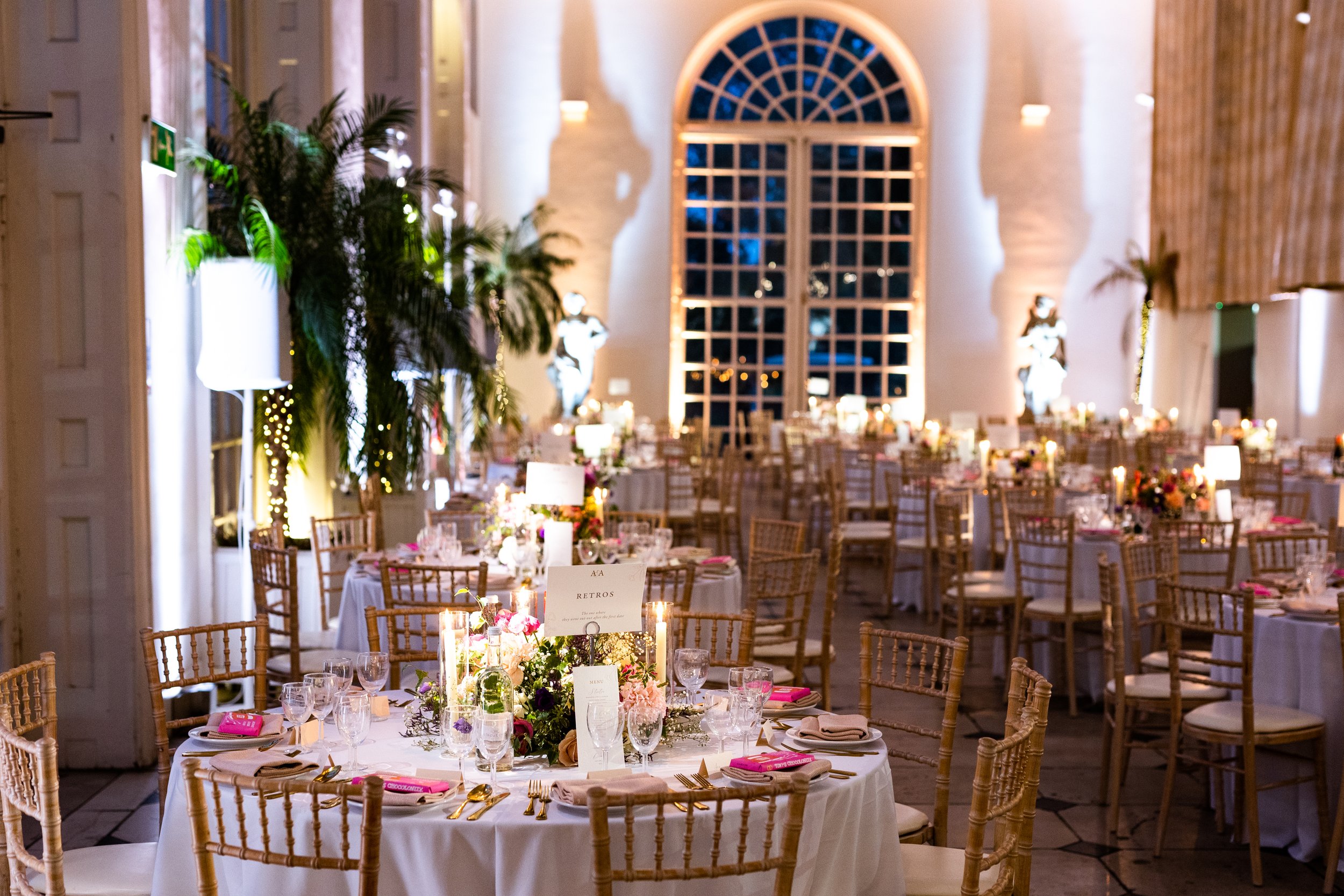 Elegant banquet hall decorated with round tables set with white tablecloths, pink napkins, glassware, and gold utensils. Floral centerpieces, tall candles, and table signs adorn each table. Indoor setting with large arched window, statues, wall light