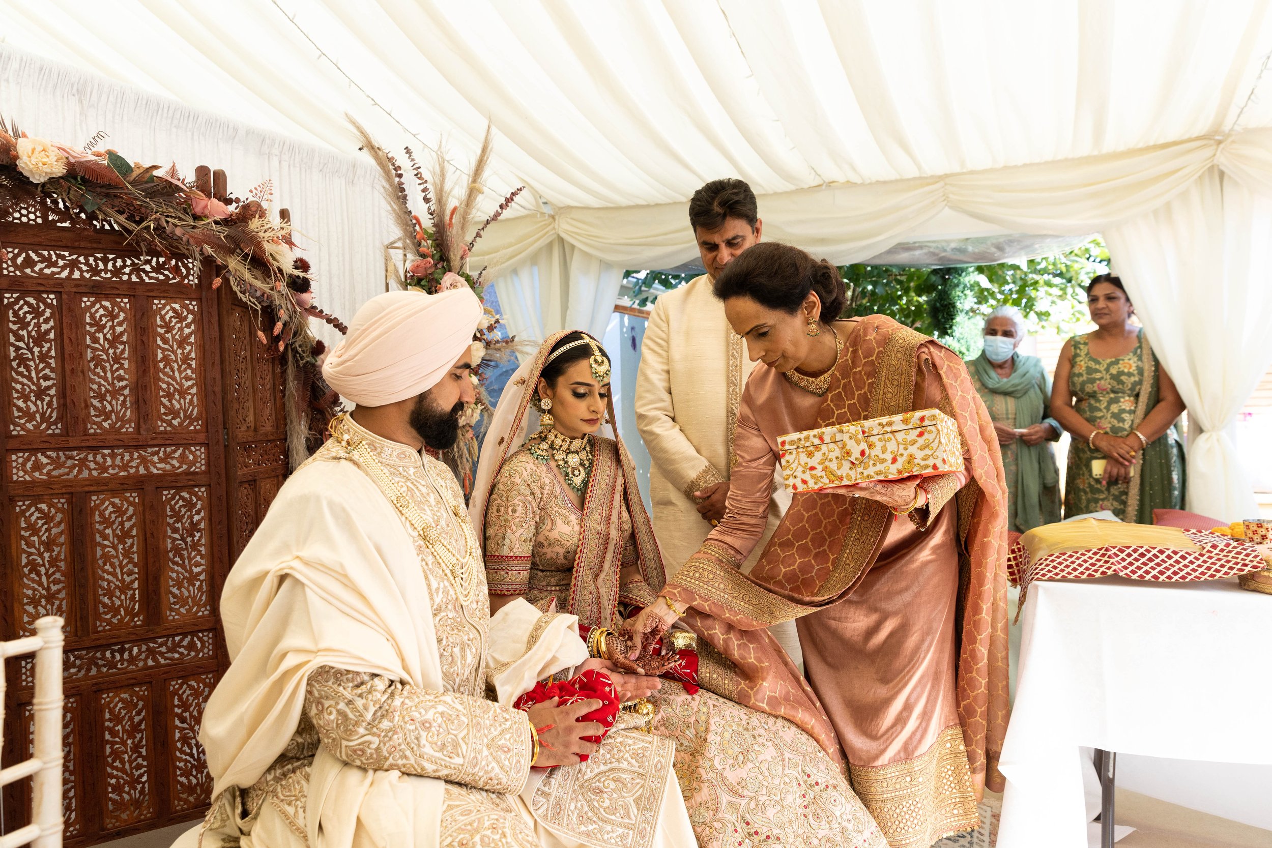 Indian wedding ceremony with bride and groom seated, woman placing a ring, and guests observing under a white canopy.