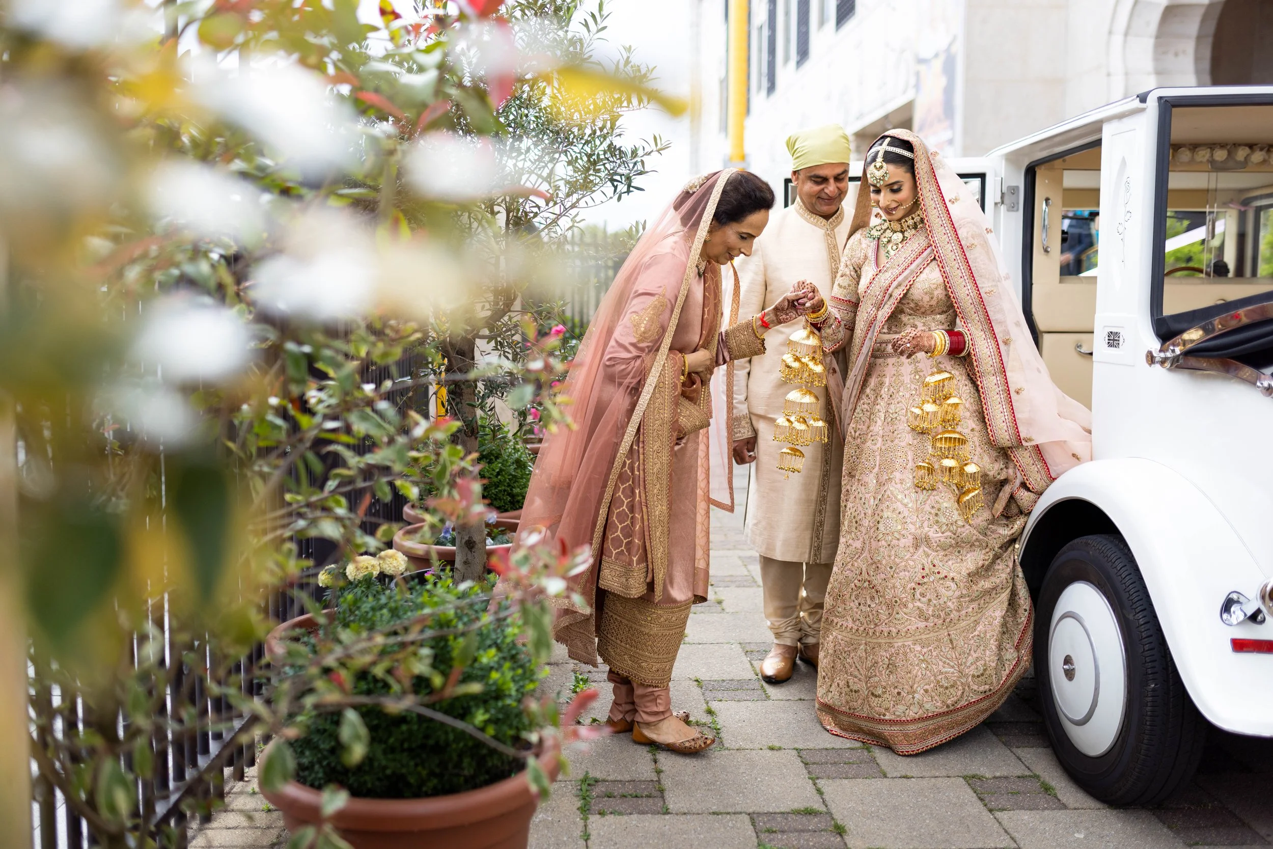 Indian bride in bridal attire getting out of a white car with gold decorations, assisted by family members during a wedding celebration.