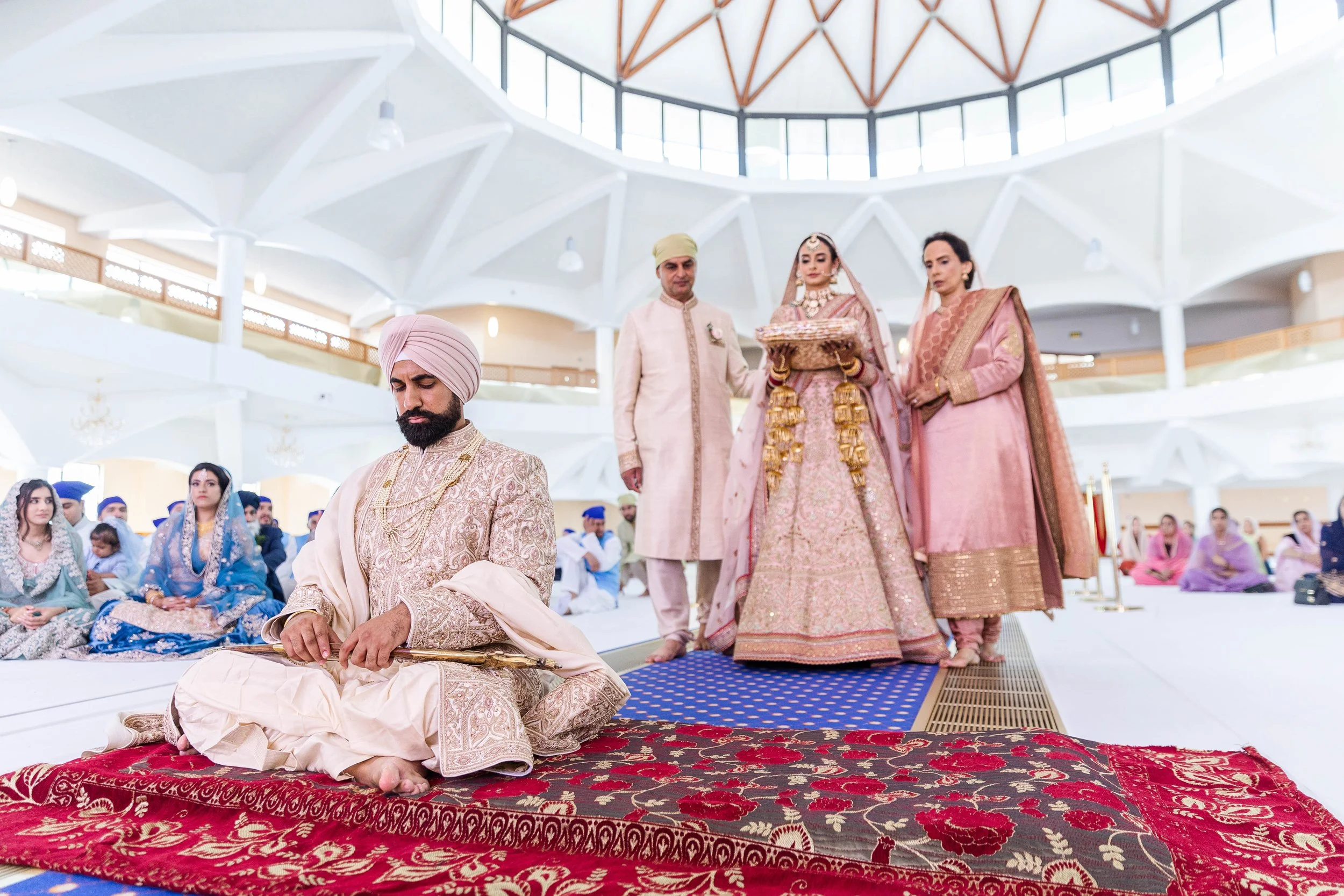 Indian wedding ceremony with groom kneeling on a decorative carpet, surrounded by family and friends in traditional attire, inside a modern venue with a domed ceiling.