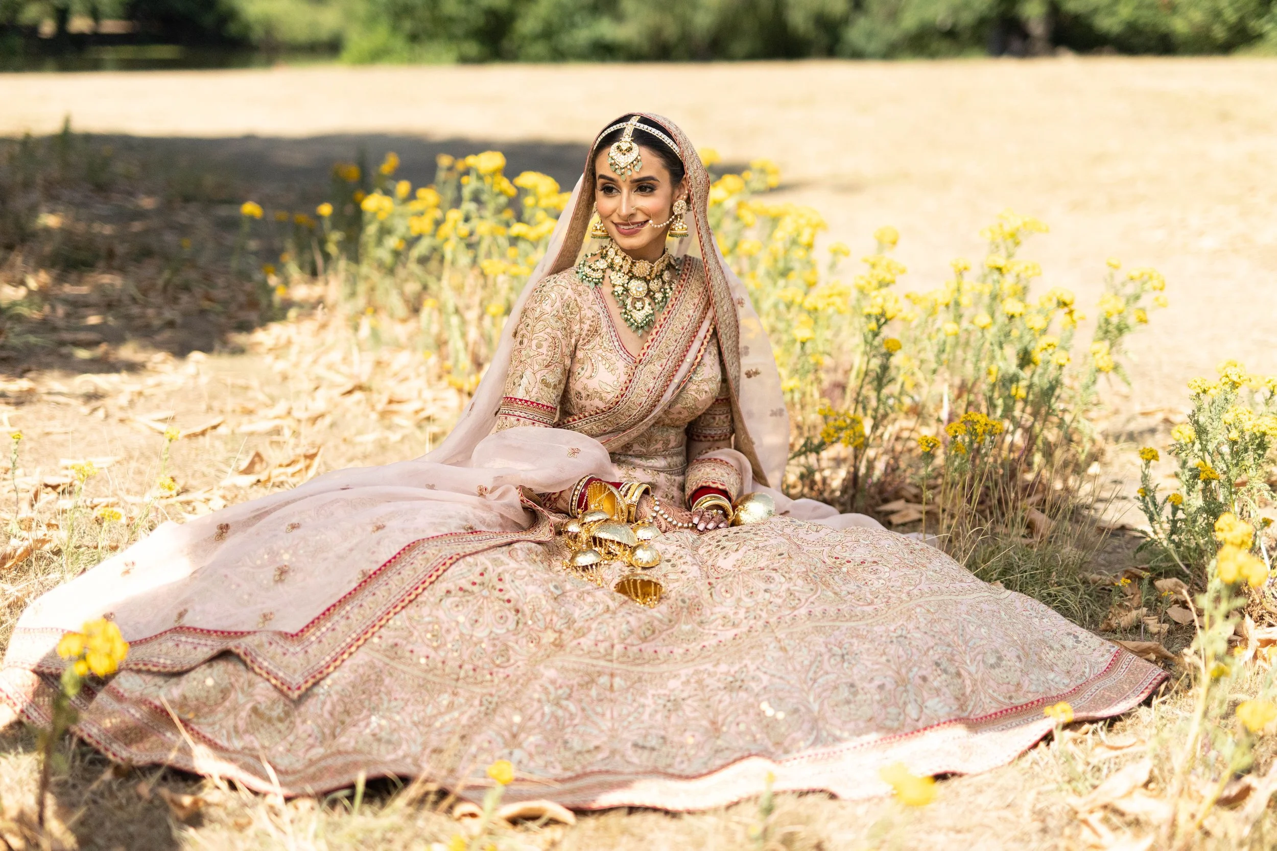 A woman dressed in traditional Indian bridal attire, sitting outdoors on the ground surrounded by yellow flowers.