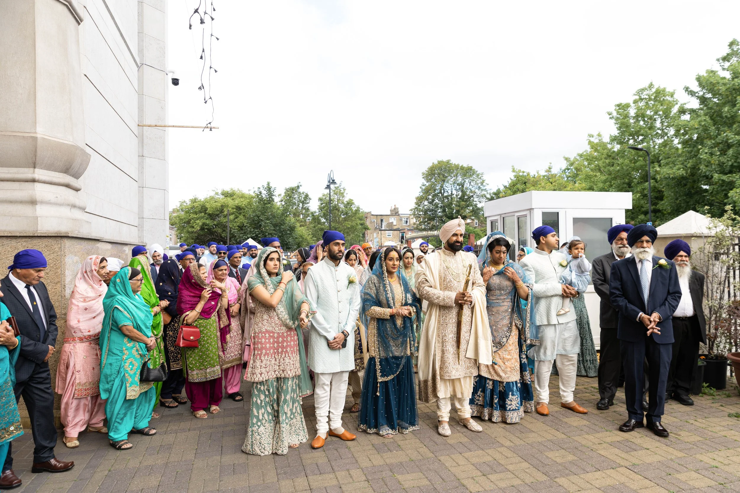 A diverse group of people dressed in traditional South Asian attire gathered outdoors for a wedding ceremony, some wearing turbans and saris, standing in front of a building with trees in the background.