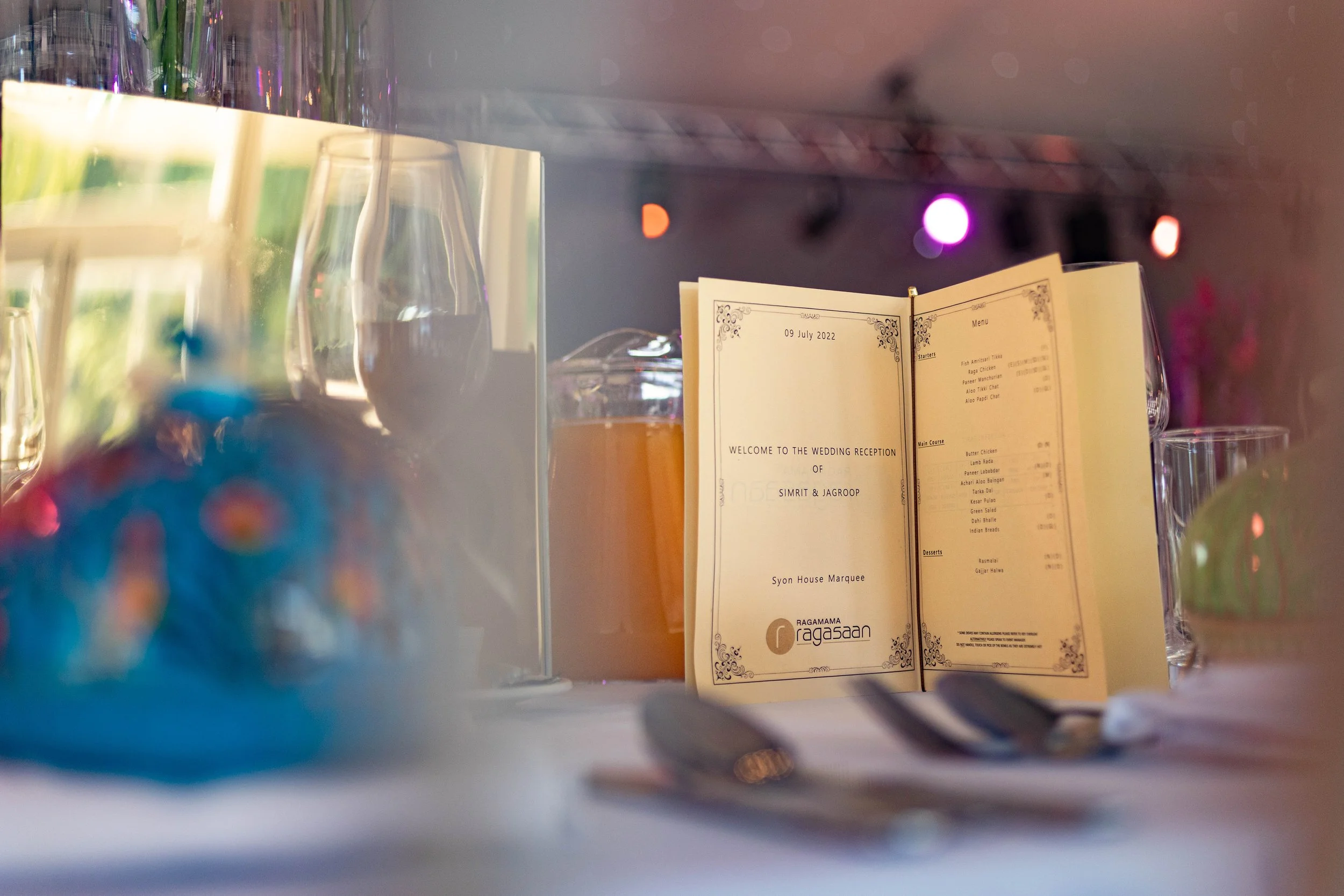 A wedding reception table setup with a fork and knife in the foreground, a menu on a stand, glasses, and decorative elements in the background.