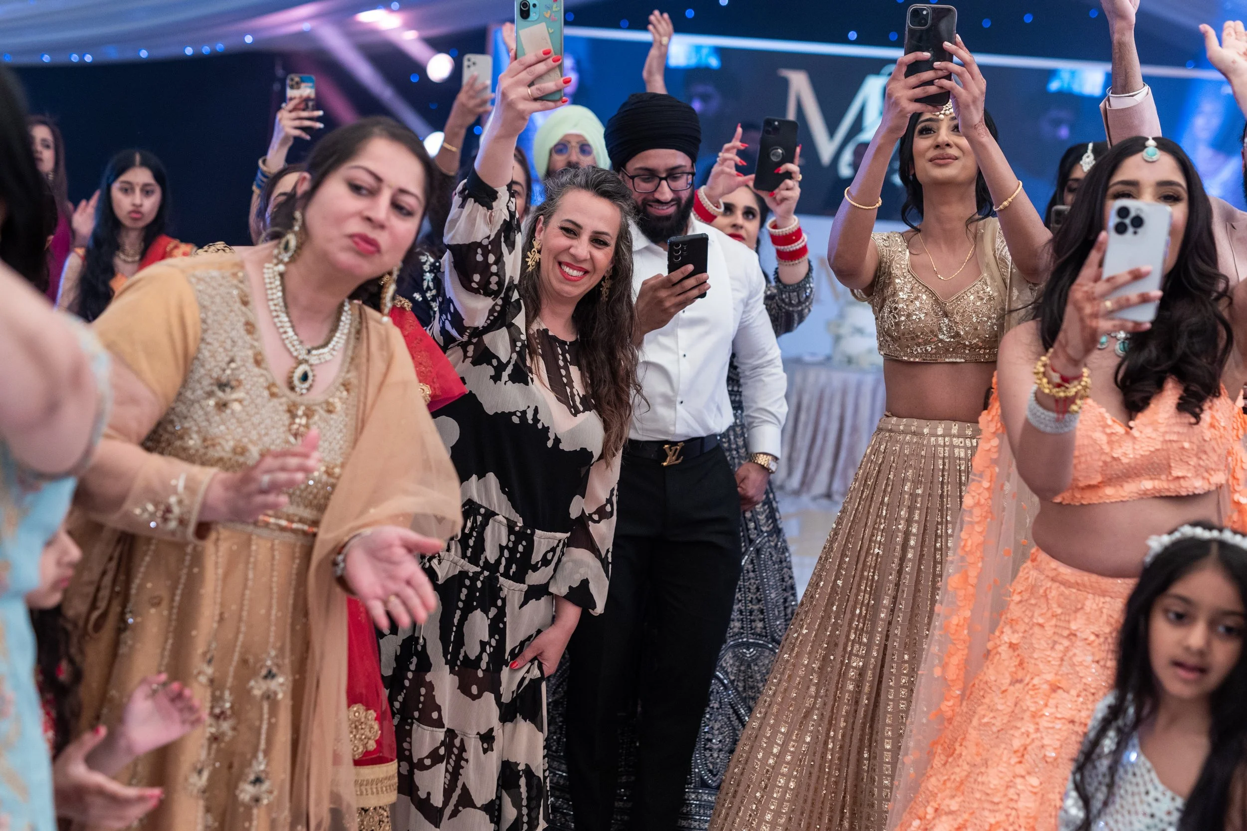 Group of people at a celebration, some taking photos or videos with smartphones, some dancing or clapping, dressed in traditional and festive clothing.