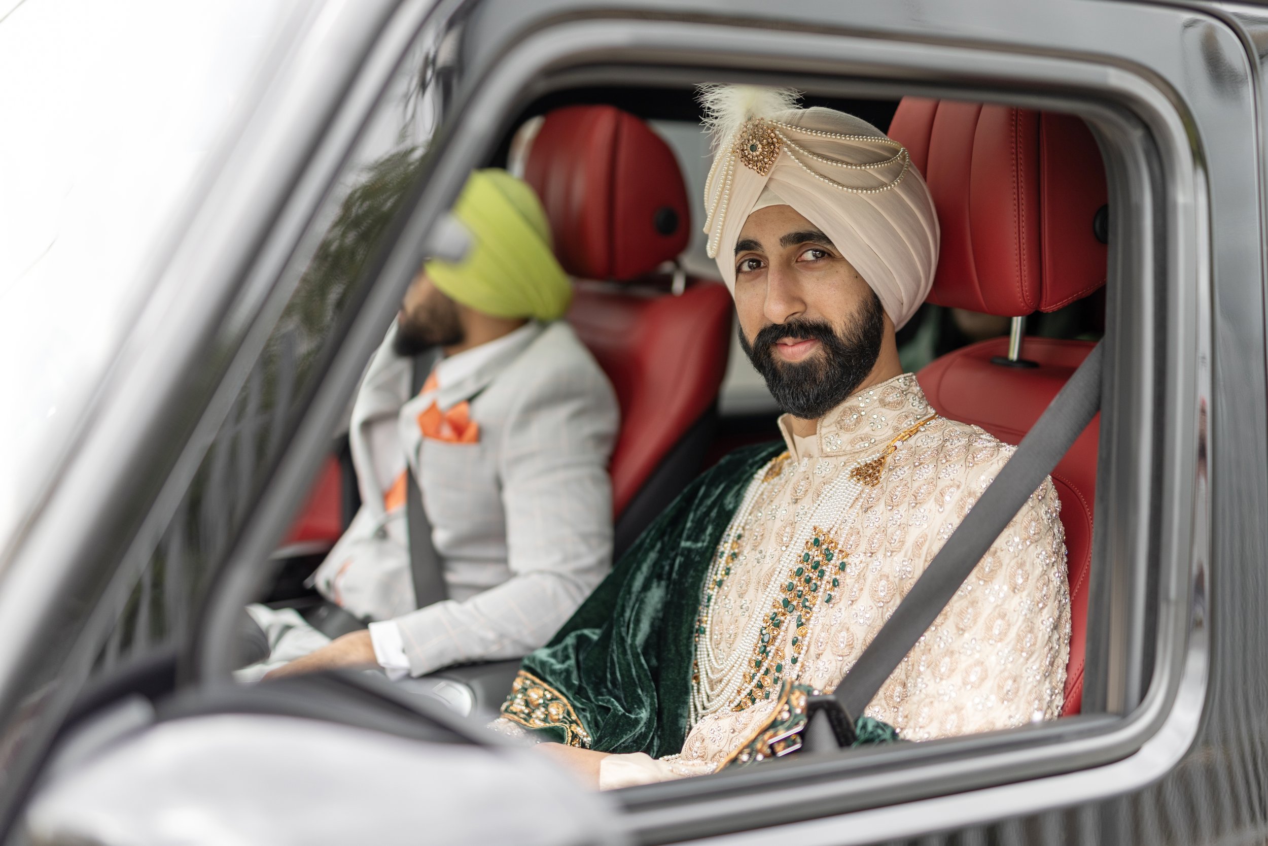 A man dressed in traditional Indian wedding attire, including a turban and ornate sherwani, sitting in a car with red leather seats alongside another man in a suit with a turban.