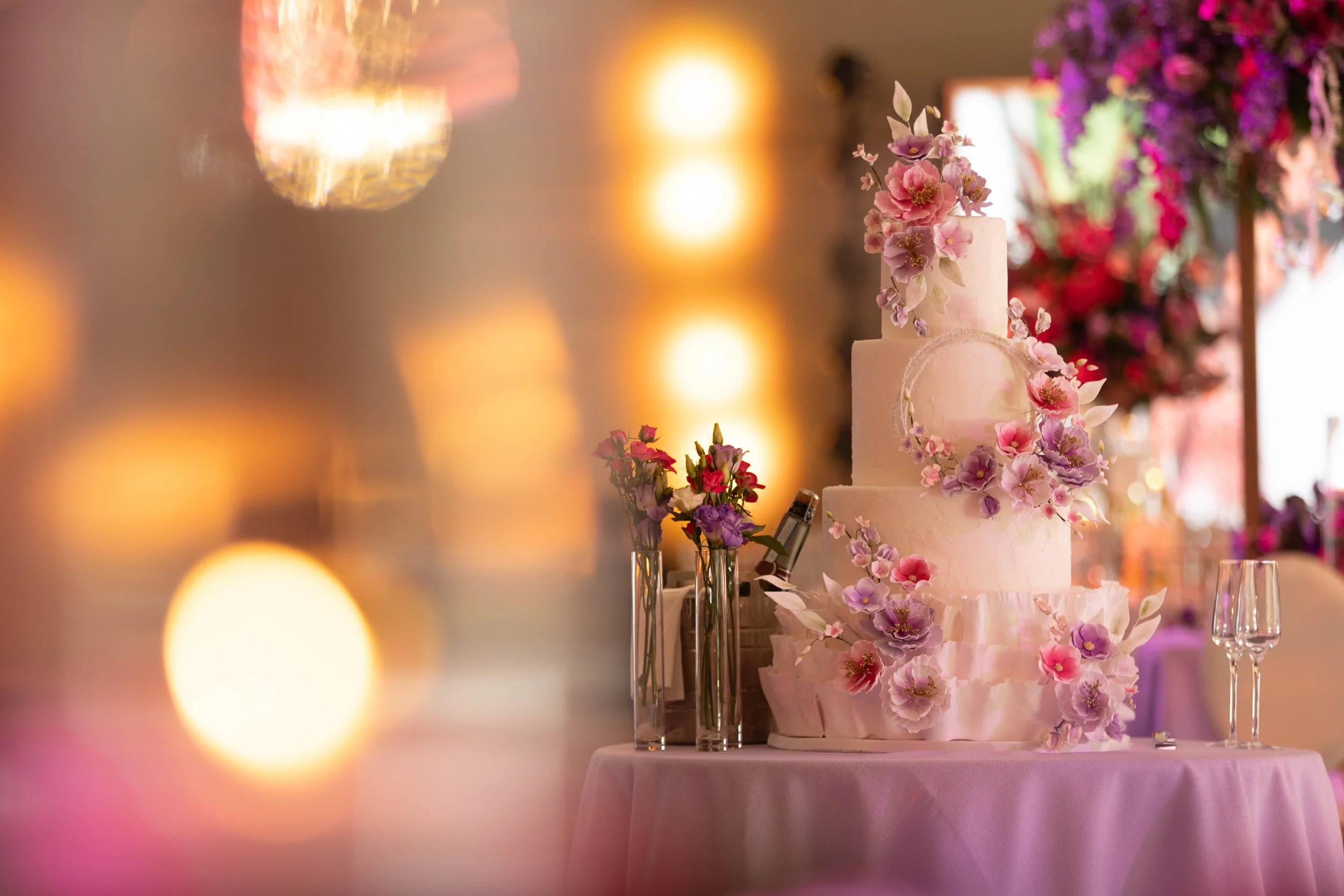 A multi-tiered wedding cake decorated with pink and purple flowers on a pink tablecloth, with blurred colorful lights in the background.