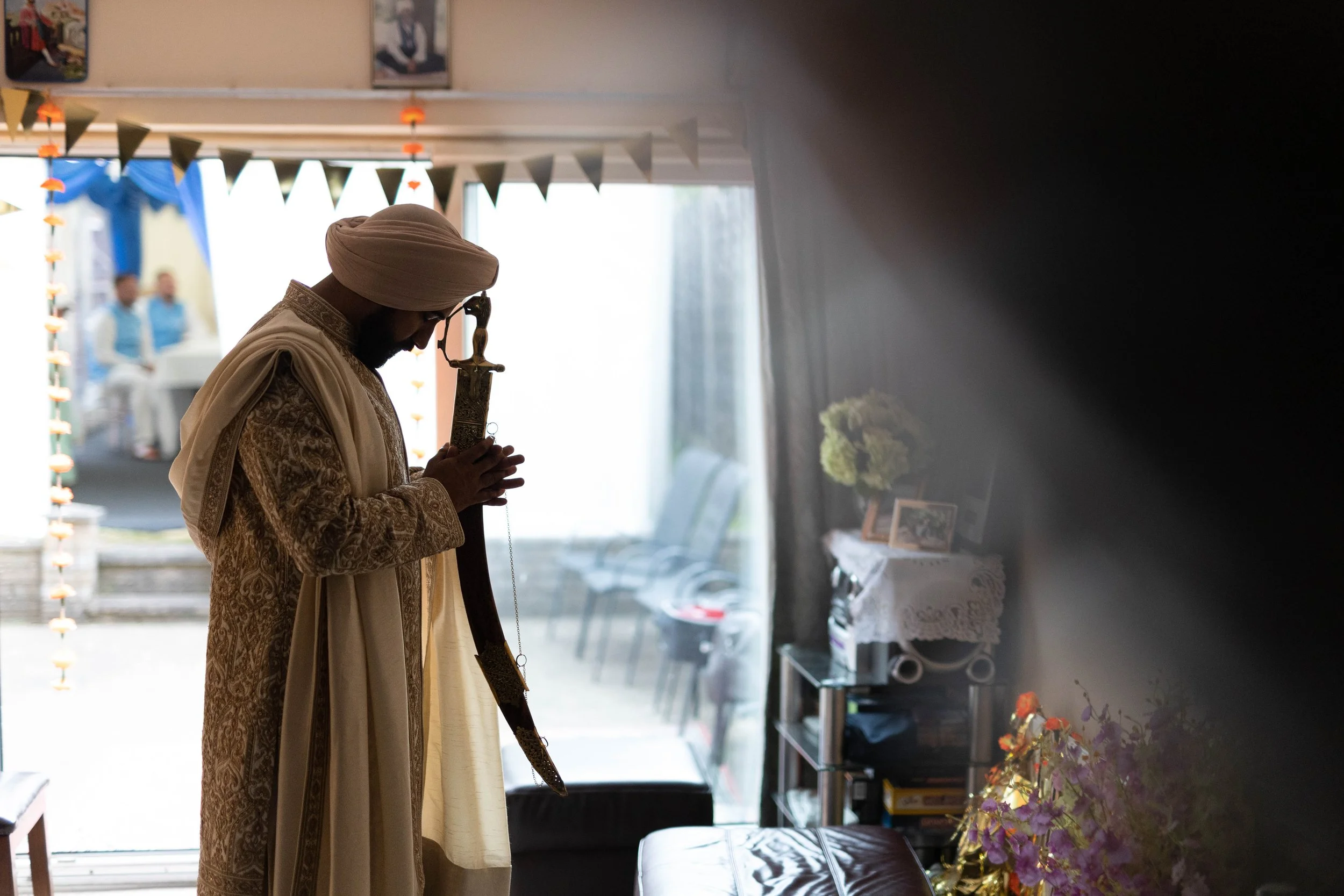 A man dressed in traditional beige clothing and a turban, standing with his hands clasped in prayer and holding a sword, inside a room with sunlight streaming in from the open door. There are chairs outside and decorative flags hanging above. Inside,