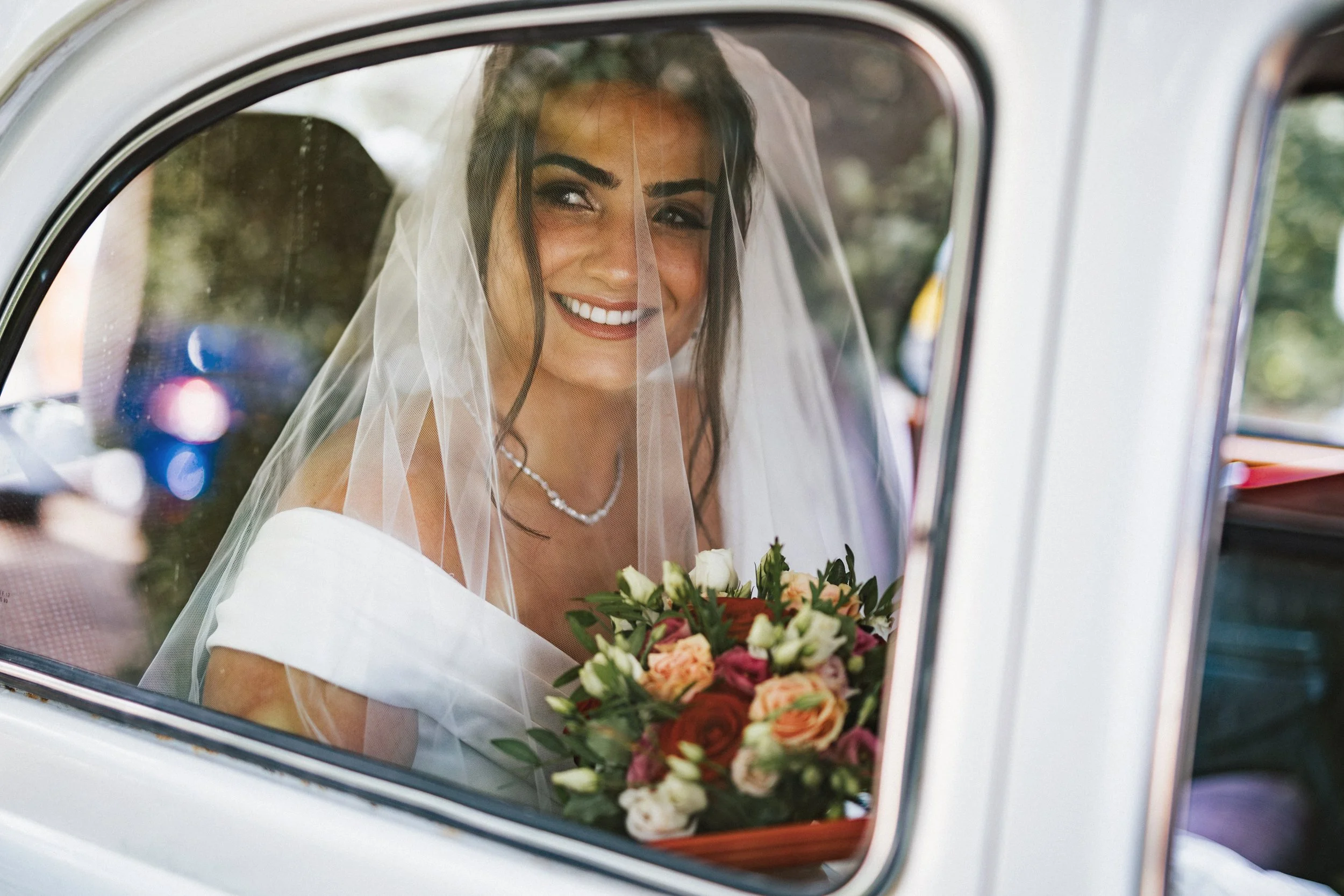 A smiling bride with dark hair and a veil, holding a bouquet of colorful flowers, sitting inside a vintage white vehicle.