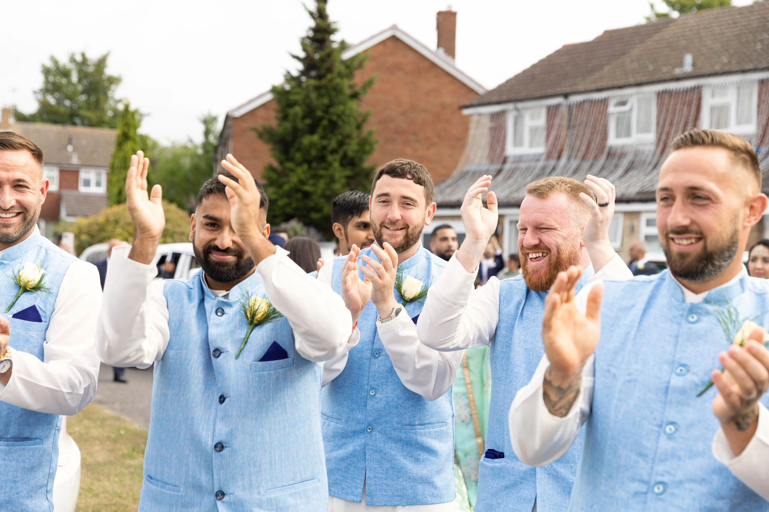 Group of men in matching blue and white traditional attire, smiling and clapping outdoors during a celebration, with houses and trees in the background.