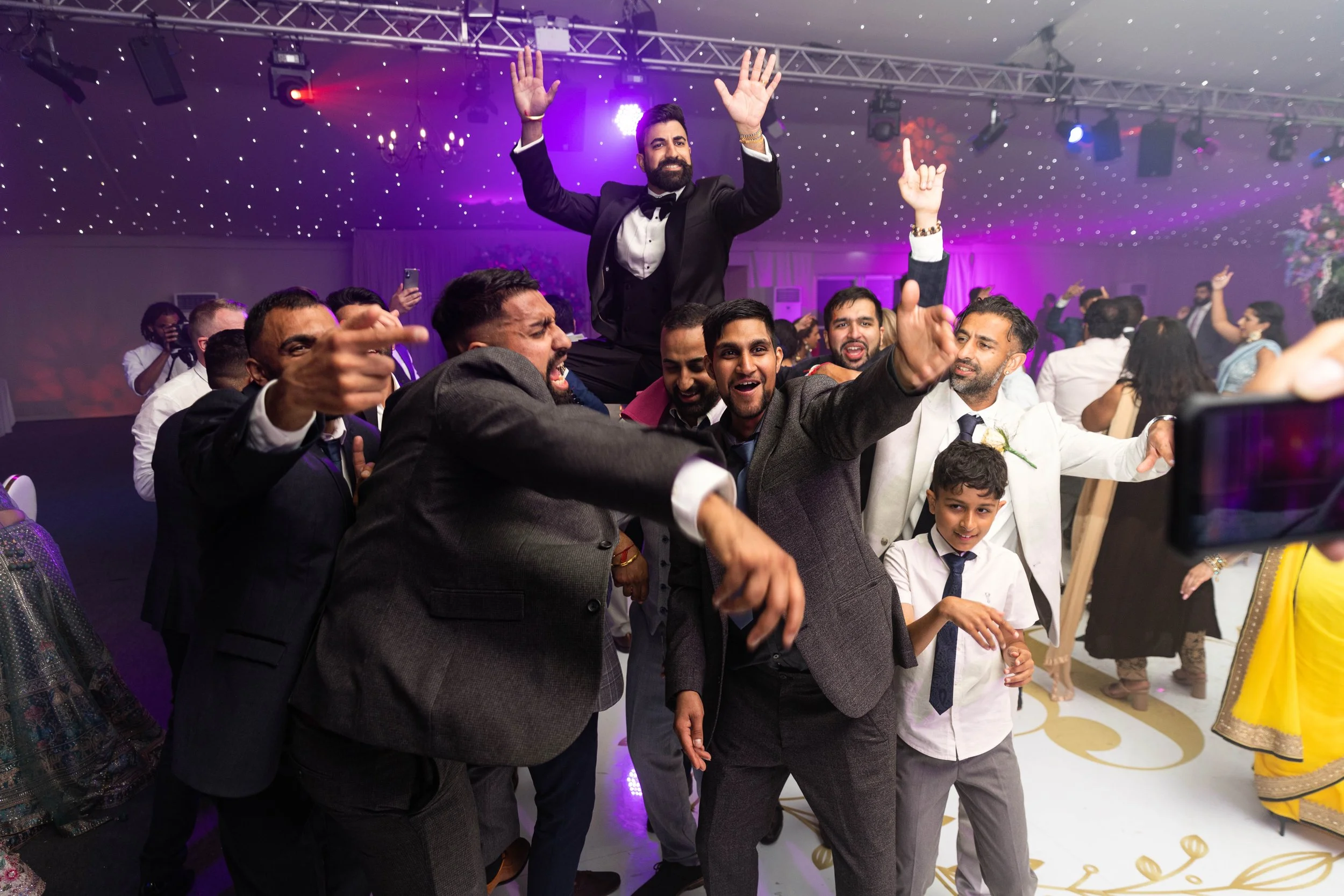 People celebrating at a wedding reception, dancing and smiling in a decorated hall with purple lighting and ceiling lights.