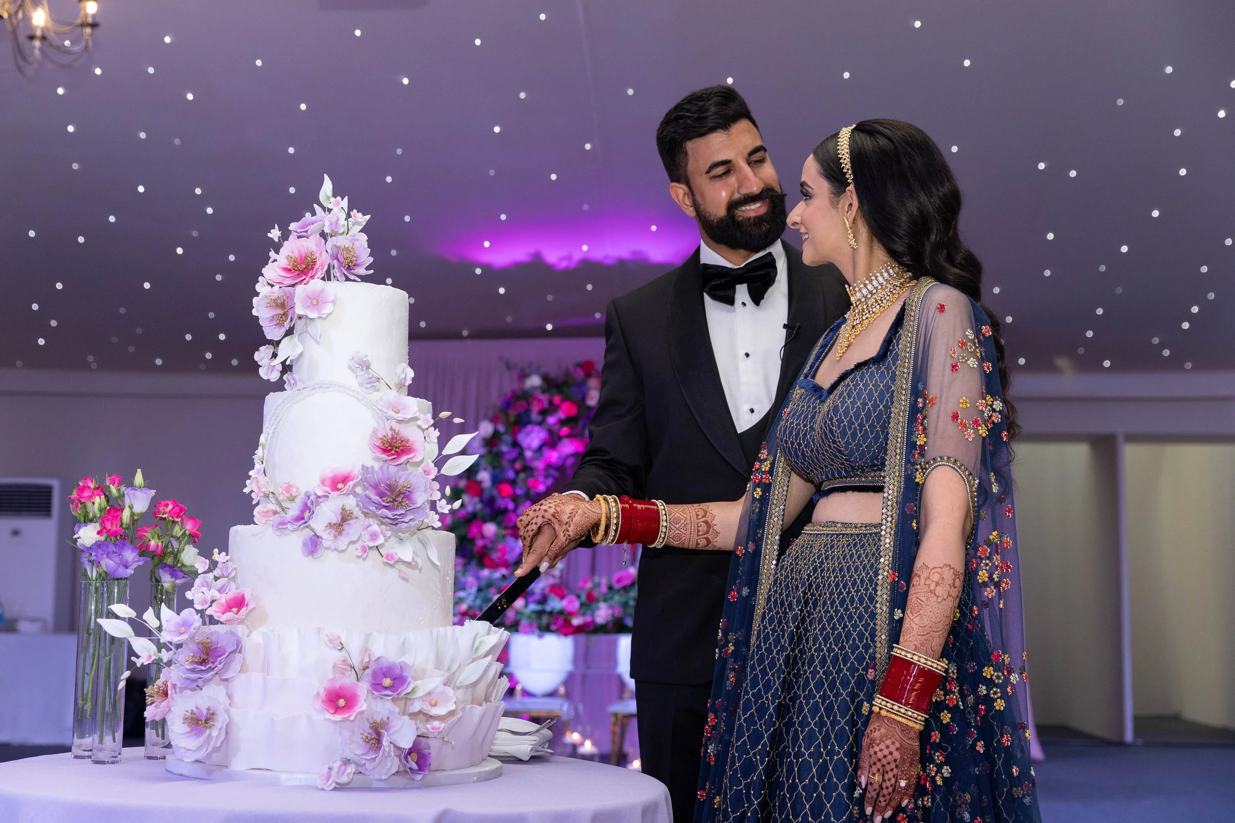A newlywed Indian couple in traditional attire cutting a wedding cake together, surrounded by flowers, in a decorated reception hall with purple lighting and star-like ceiling lights.