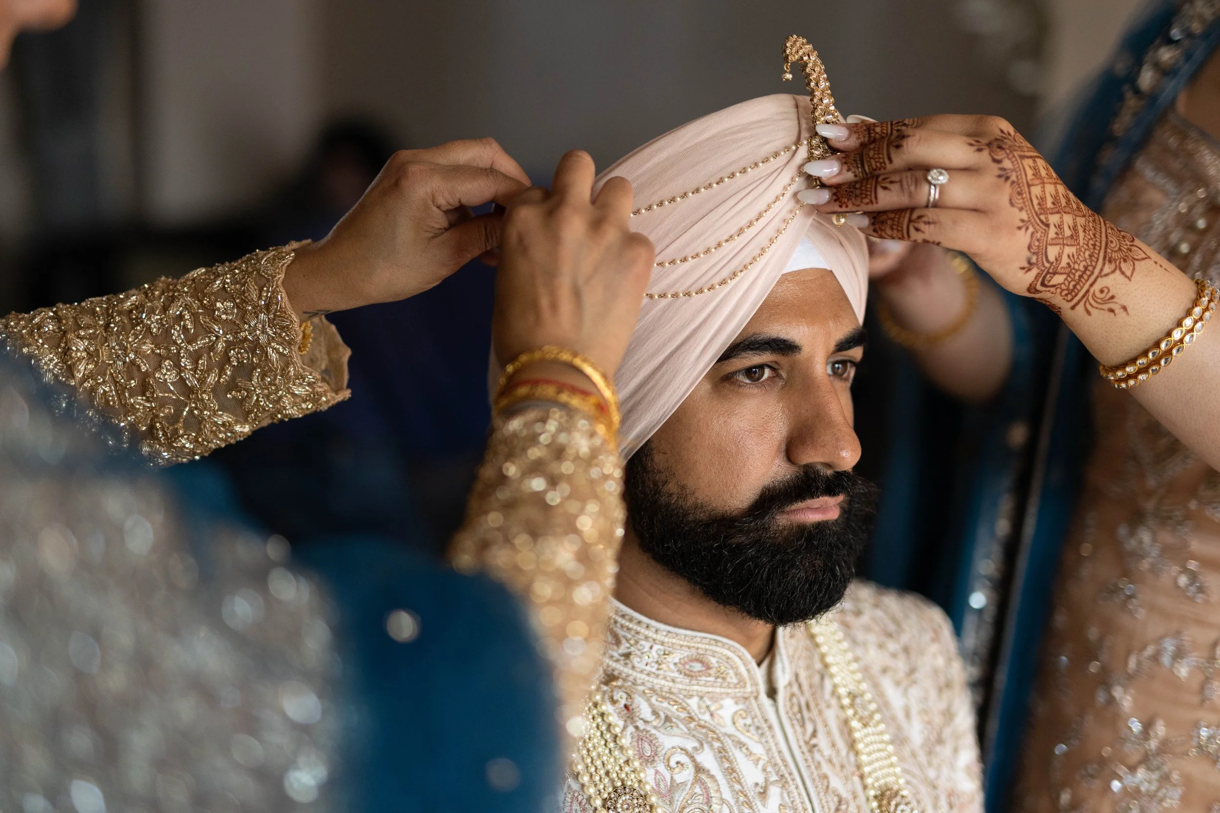 A groom with a beard and traditional attire, wearing a white turban, is being adorned with a pink turban by women dressed in ornate, embroidered sarees with jewelry, during a wedding ceremony.