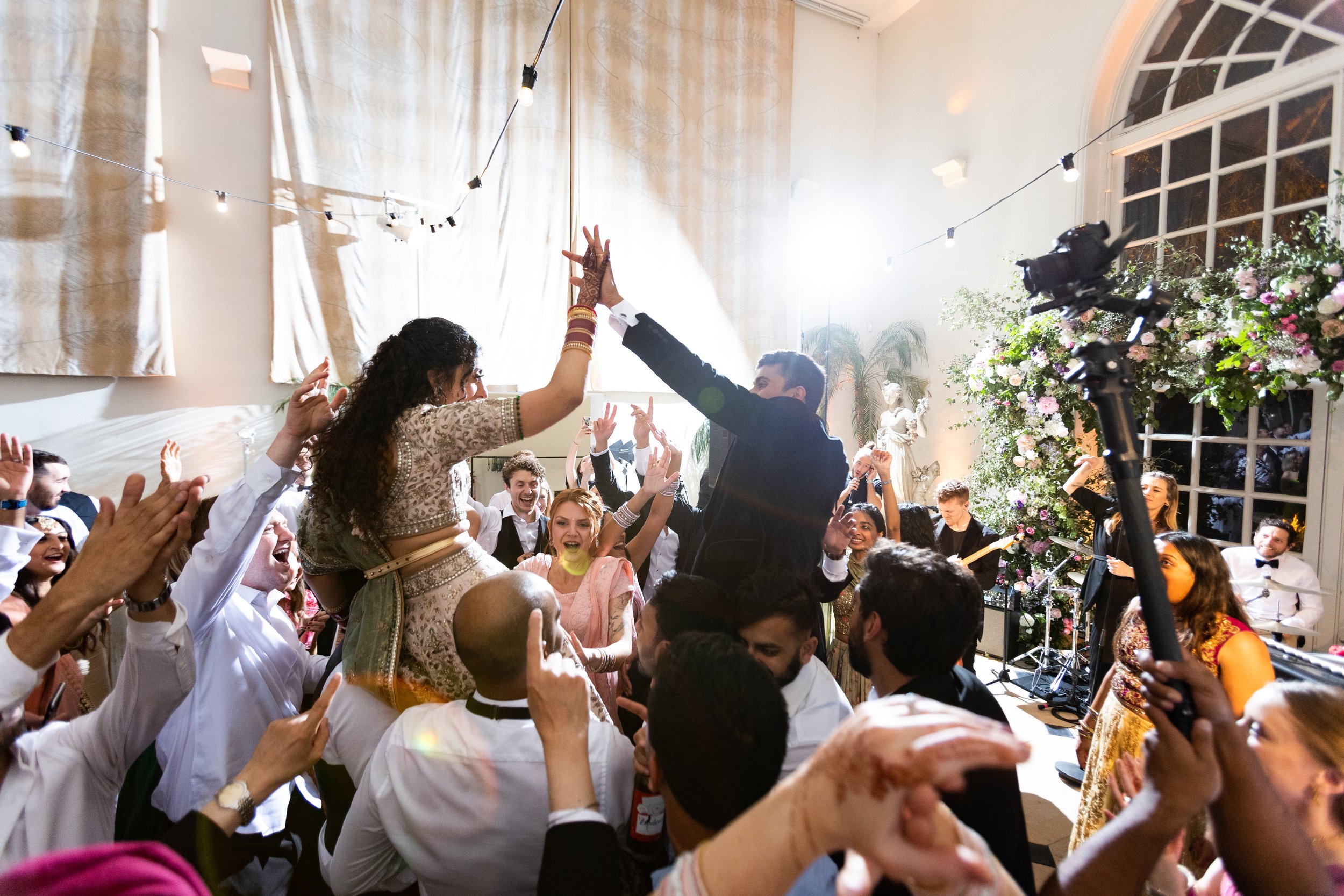 People dancing and celebrating at a lively indoor event with string lights, floral decorations, and a band performing.