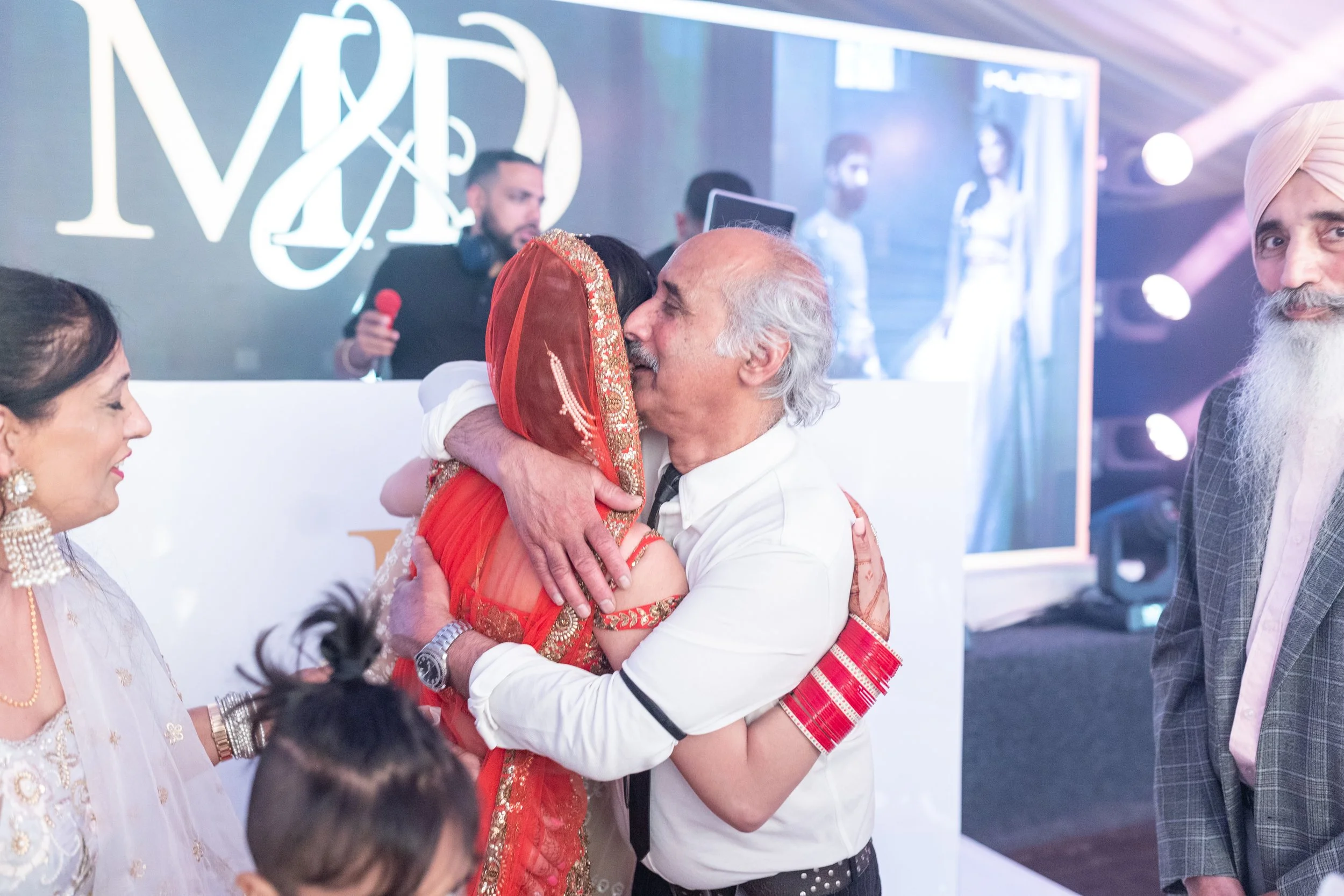 An elderly man hugging a woman in traditional Indian attire at a celebration, with other women and men around and a DJ setup in the background.