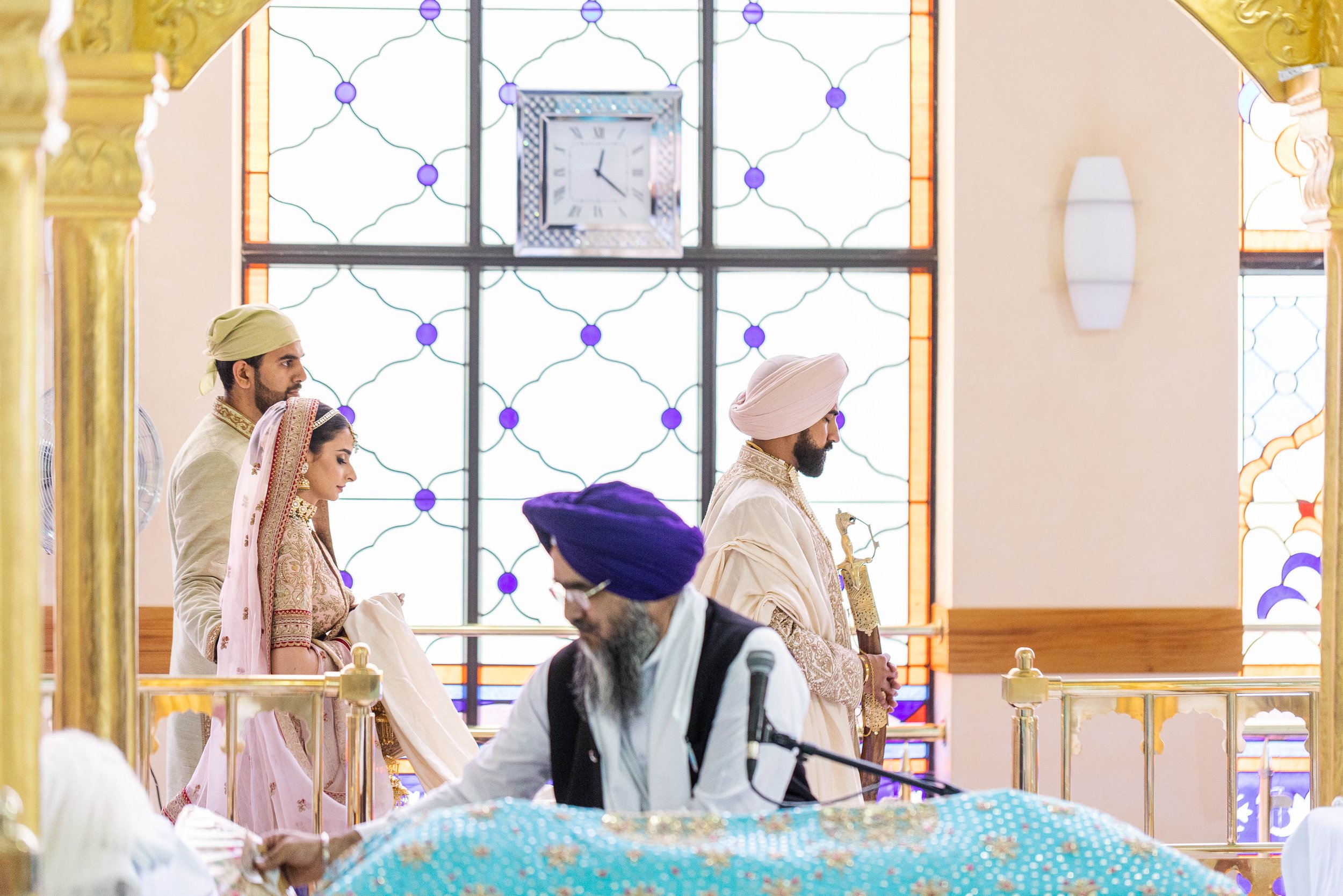 Indian wedding ceremony with the bride and groom standing before a priest, inside a decorated hall with stained glass windows and a clock.