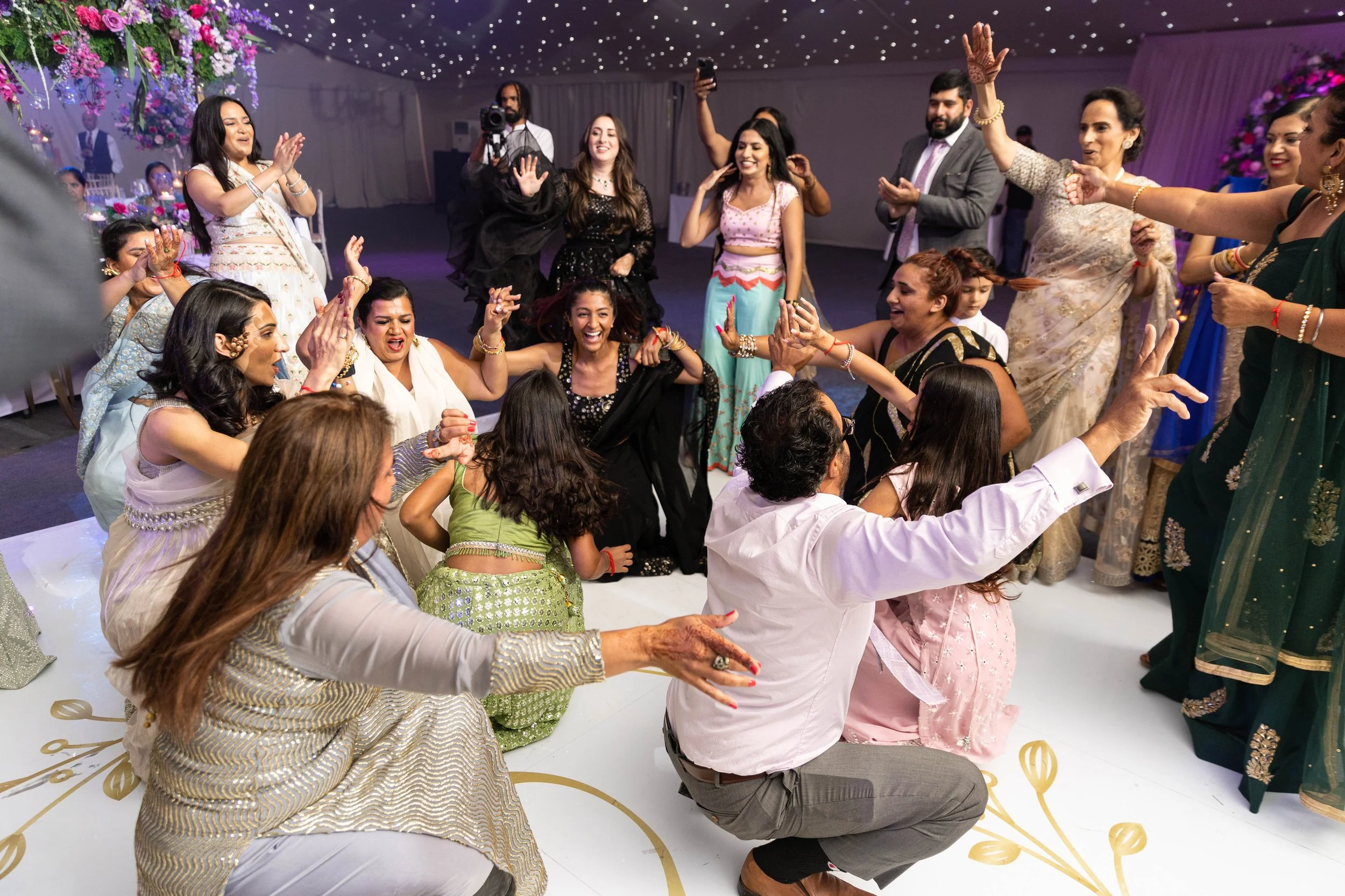 Group of people dancing and celebrating at an Indian wedding or celebration, wearing traditional sarees and outfits, with festive decorations and twinkling ceiling lights.