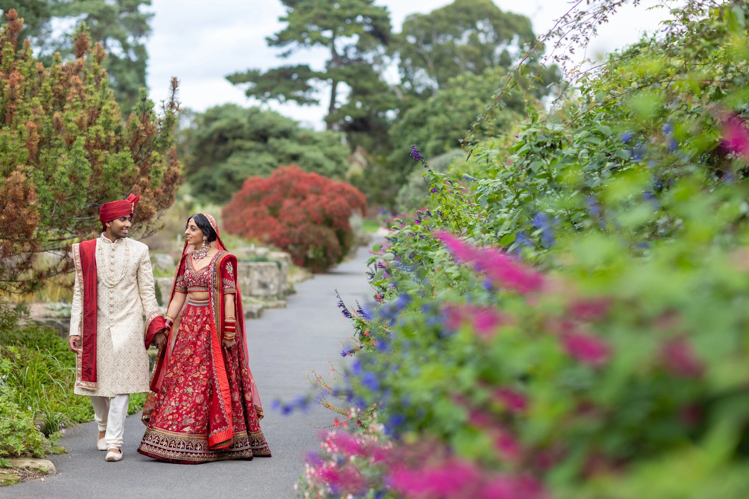 A bride and groom in traditional Indian wedding attire walking hand in hand on a garden pathway surrounded by colorful flowering bushes and trees.