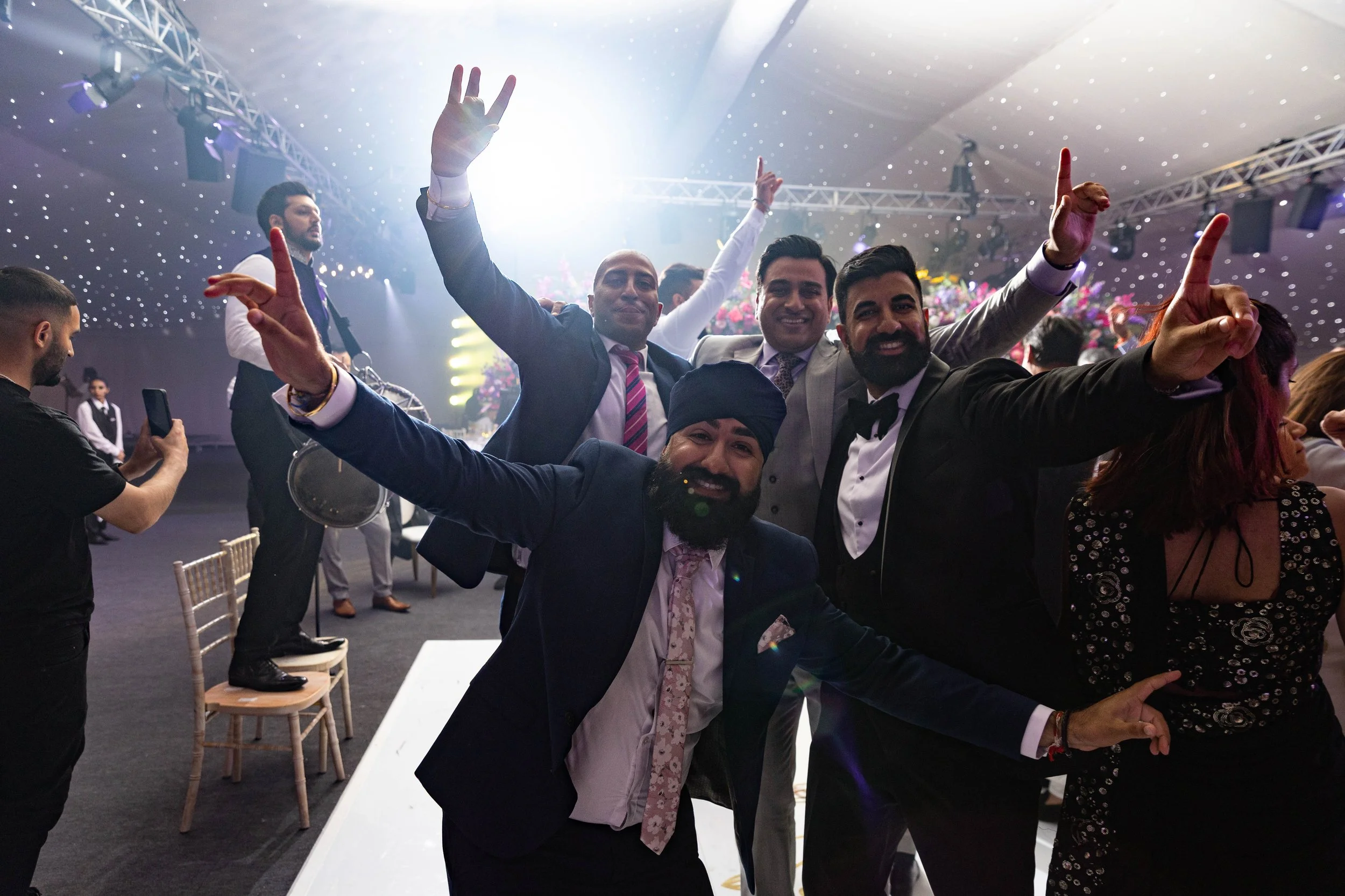 Group of men dressed in formal suits celebrating at an indoor event with a decorated ceiling and stage in the background, some standing on chairs and smiling.