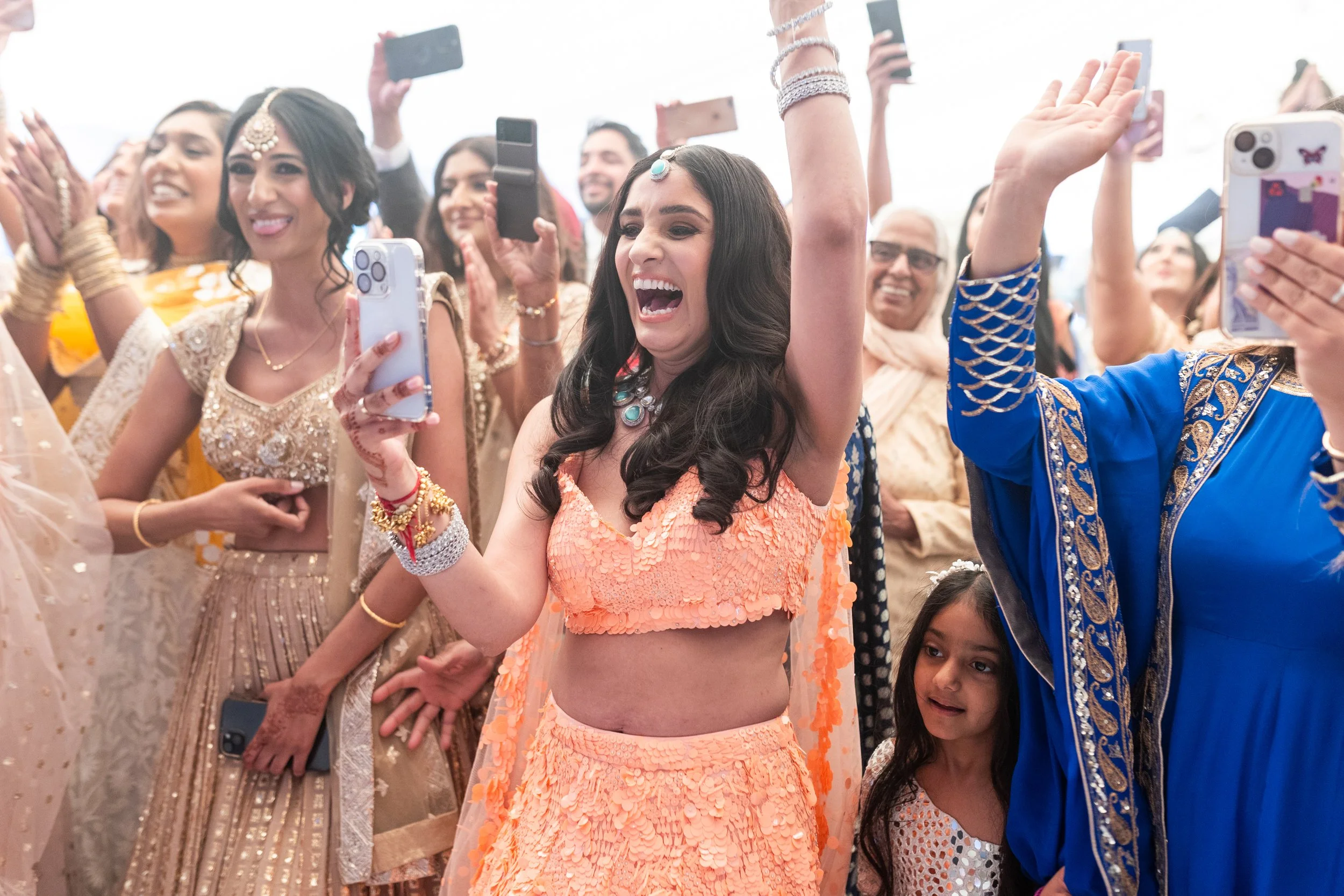 Celebration with women wearing traditional Indian attire, one woman in an orange outfit smiling and raising her arms, surrounded by others taking photos and smiling.
