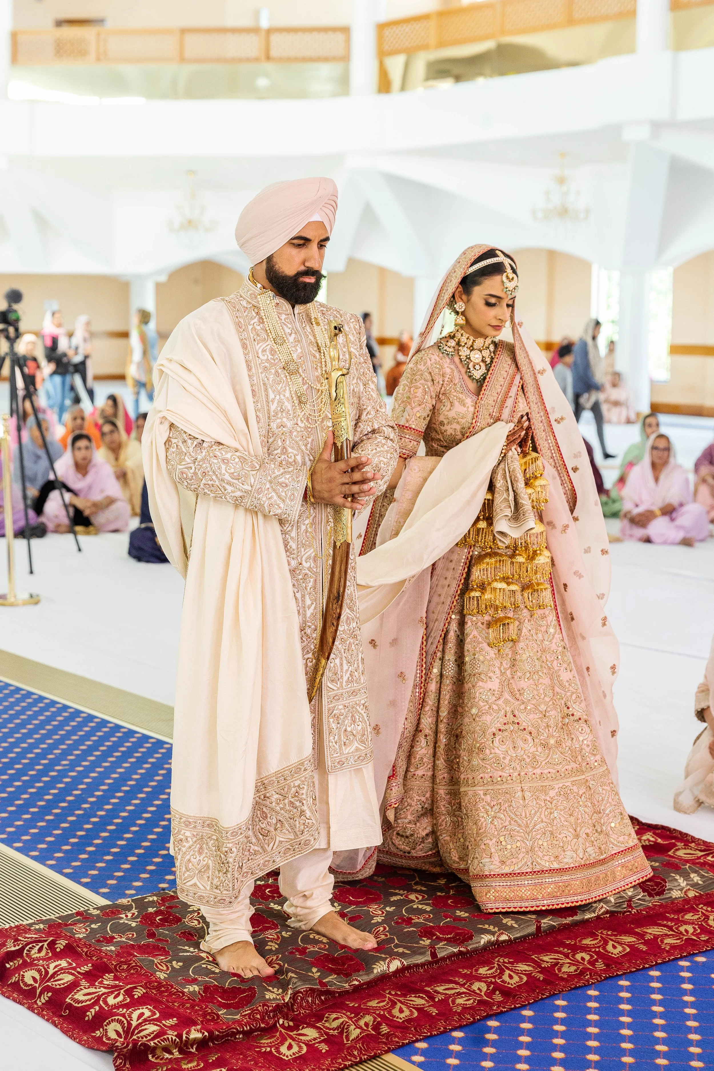 Indian bride and groom in traditional attire during a wedding ceremony, standing on a red and gold carpet with guests seated in the background.