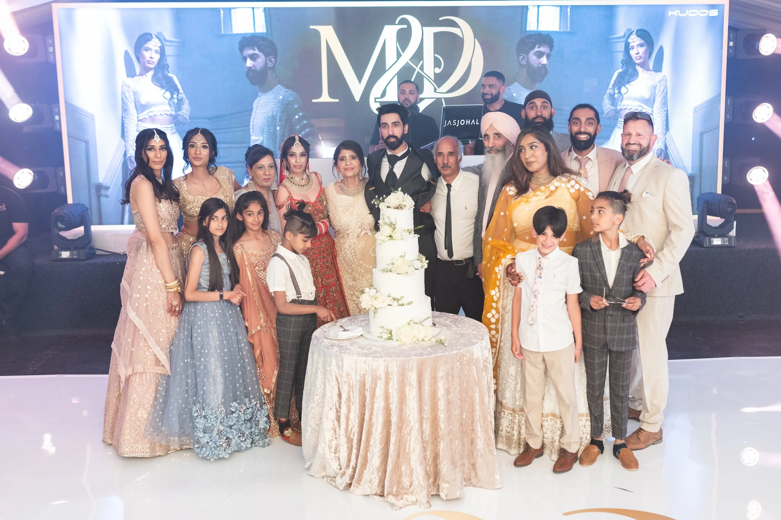 Group of people celebrating a wedding around a tall, multi-tiered white wedding cake on a table with a cream-colored tablecloth. The couple is in the center, with the bride in a white dress and the groom in a black vest and tie. The background featur