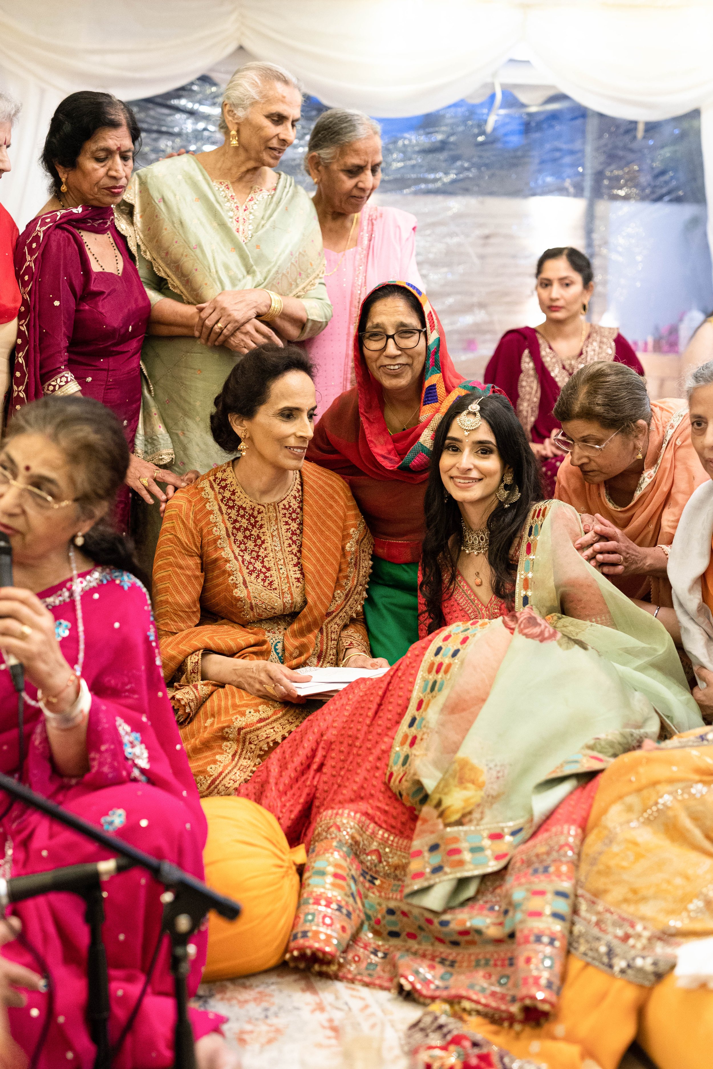 Group of women dressed in colorful traditional Indian attire, gathered around a seated woman, at a festive celebration or cultural event.