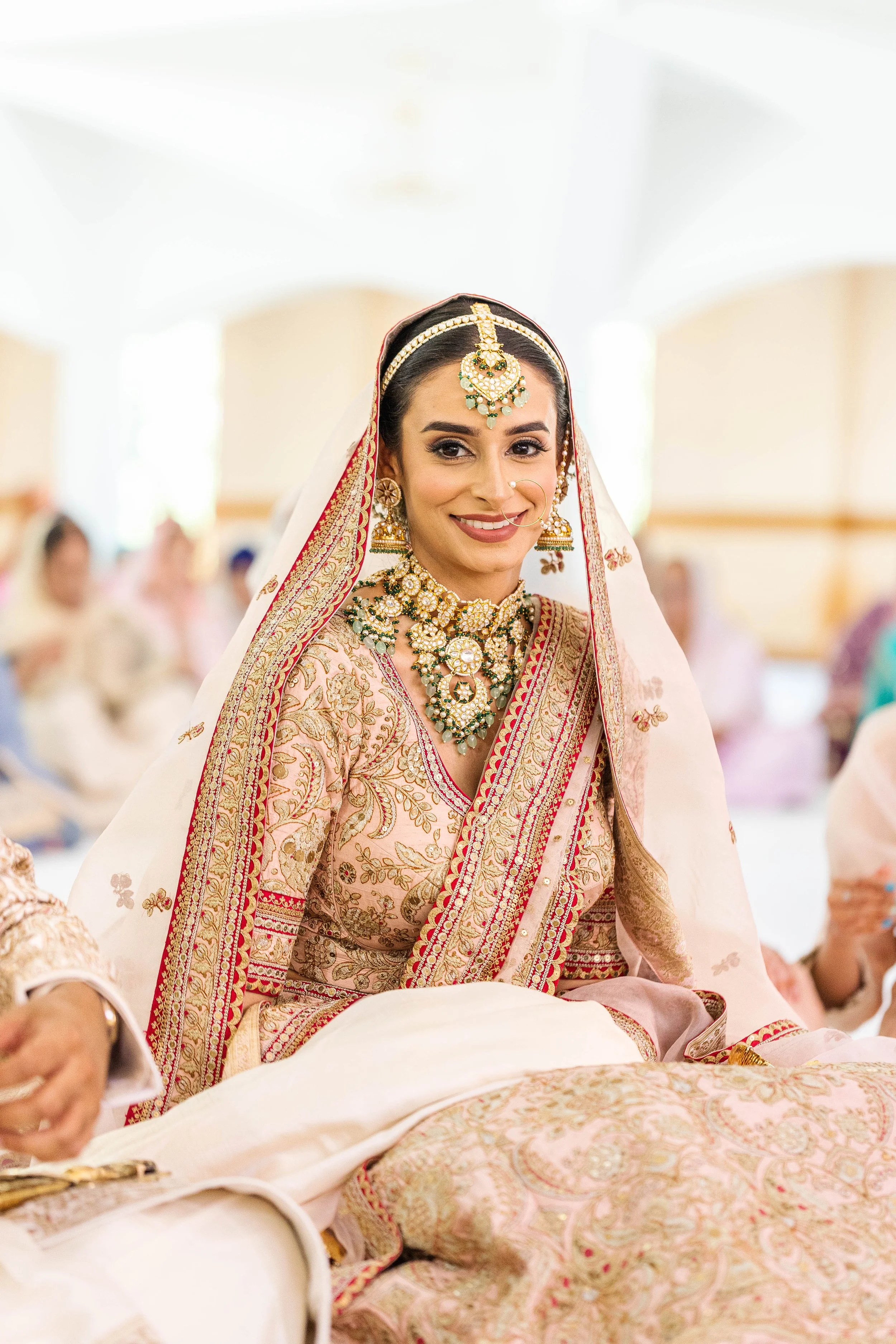 A smiling bride in traditional, ornate Indian wedding attire with jewelry, seated indoors among other guests.