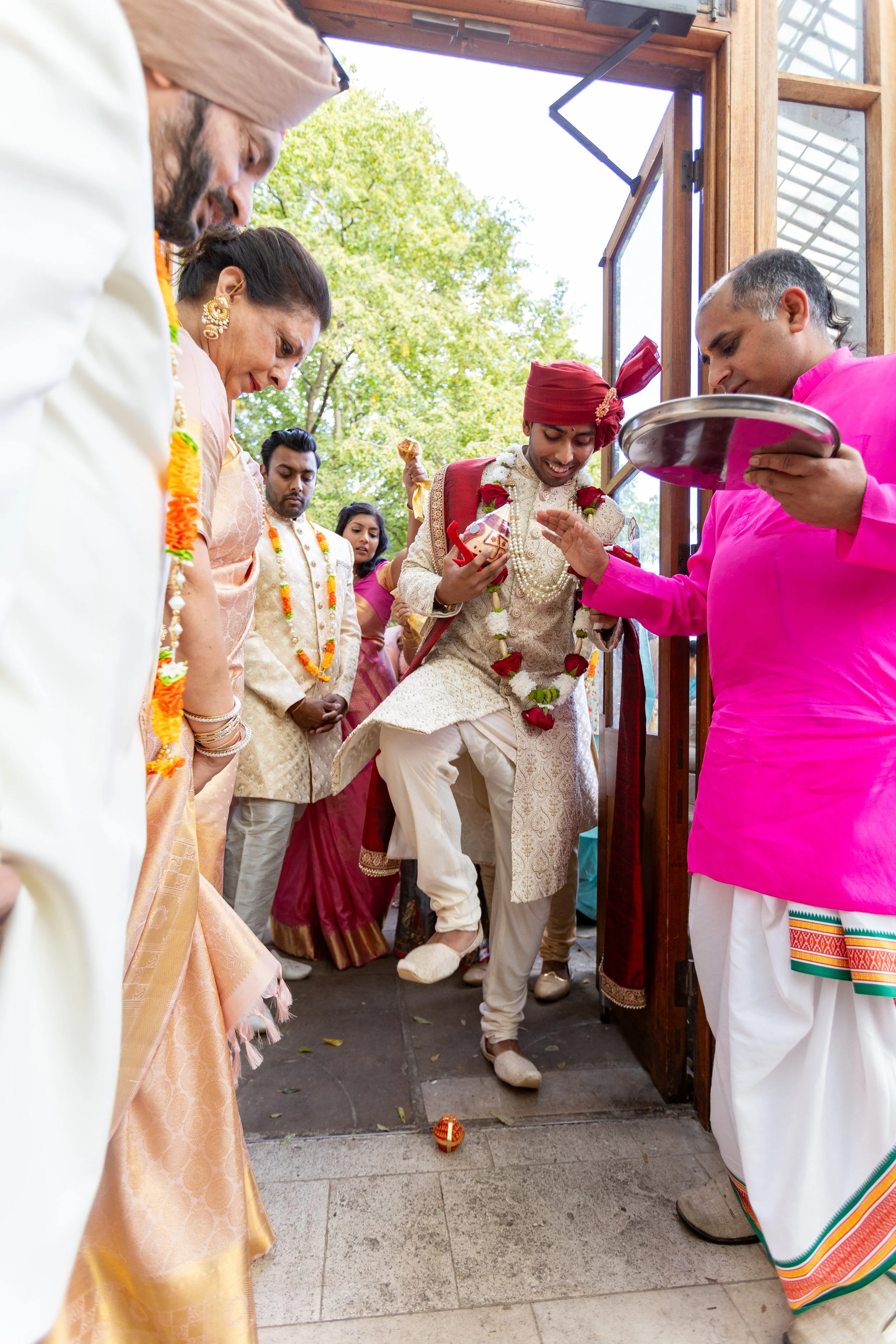 Hindu wedding ceremony with a groom stepping over a lit traditional fire while family members stand around, dressed in colorful attire.