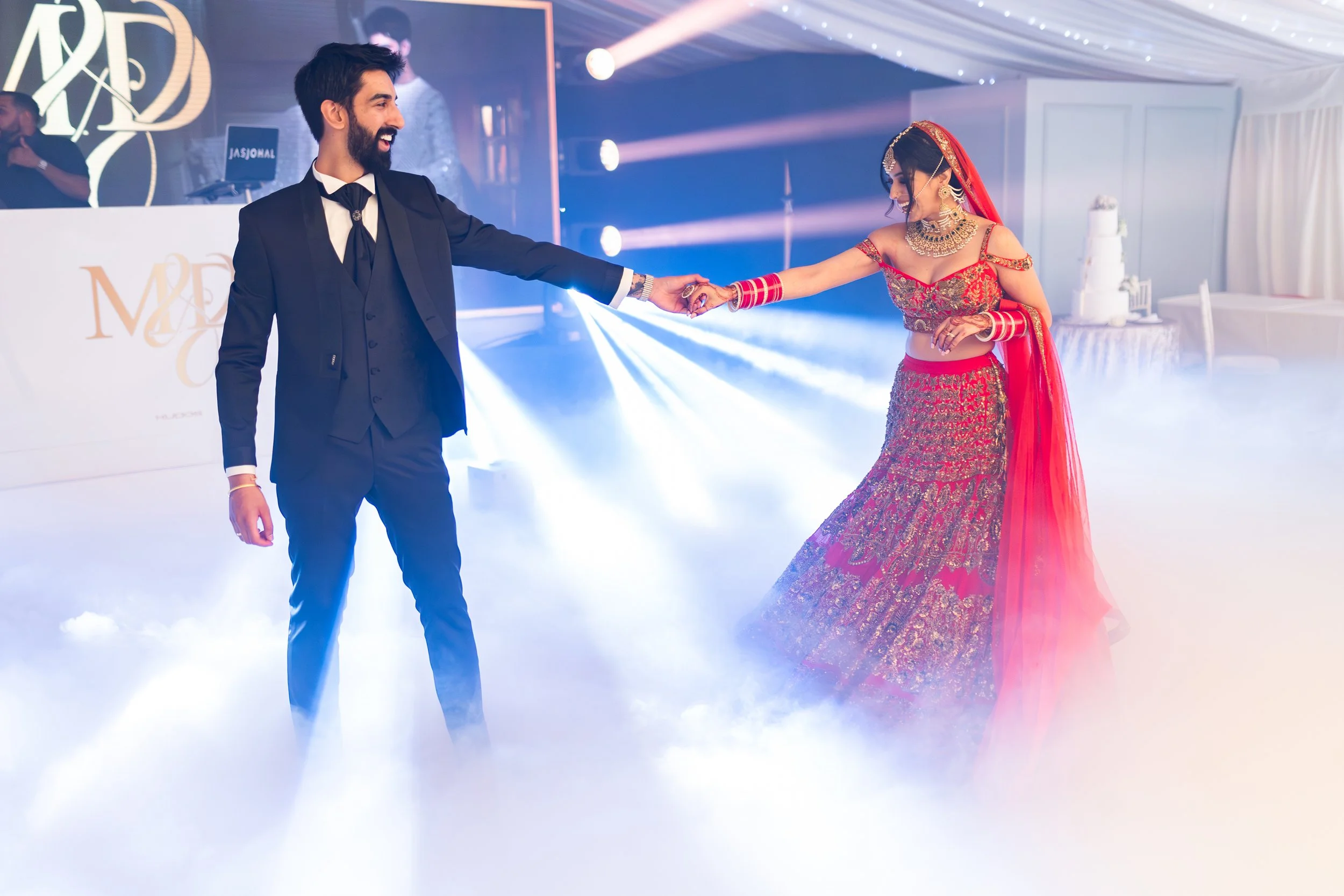 A bride and groom dancing at their wedding reception. The groom is wearing a black suit and tie, and the bride is dressed in a red traditional Indian bridal outfit with jewelry and bangles. They are holding hands, smiling, and dancing in a decorated 