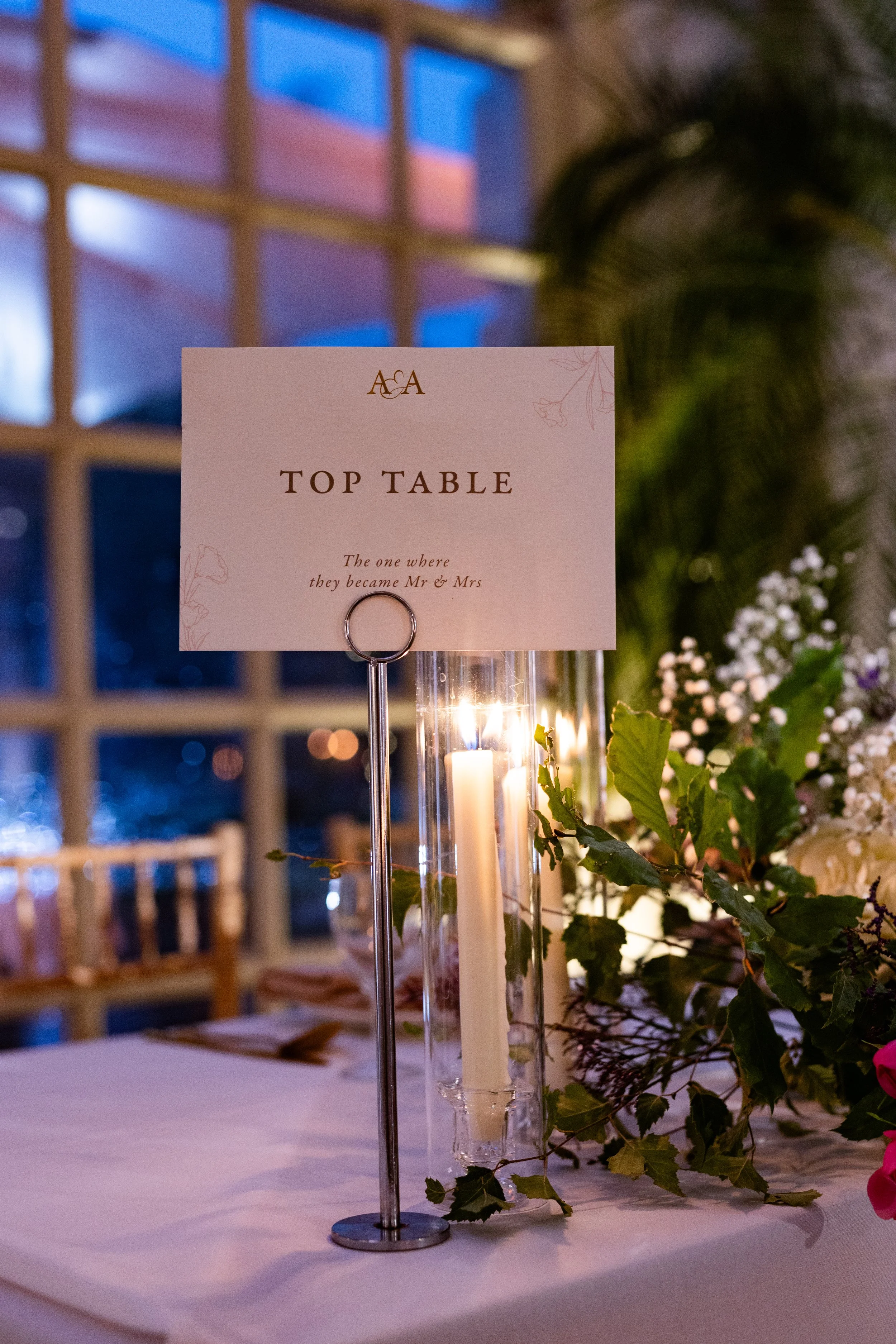Wedding reception table sign indicating the "Top Table" with a candle and floral decorations, and a window in the background.