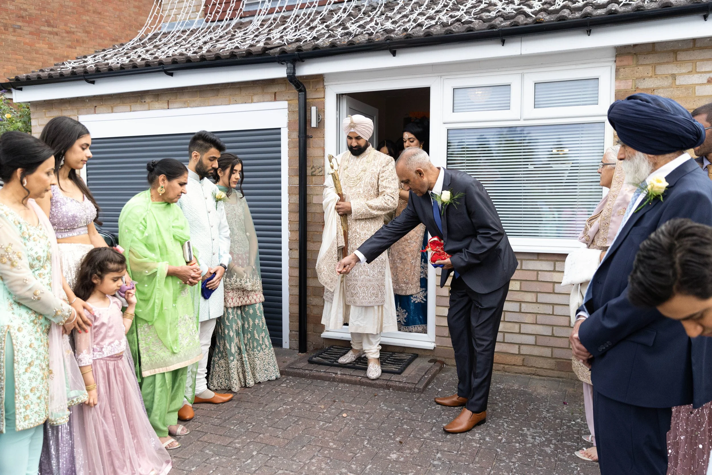 People participating in a traditional Indian housewarming ceremony outside a brick house, with one man pouring water as others observe.