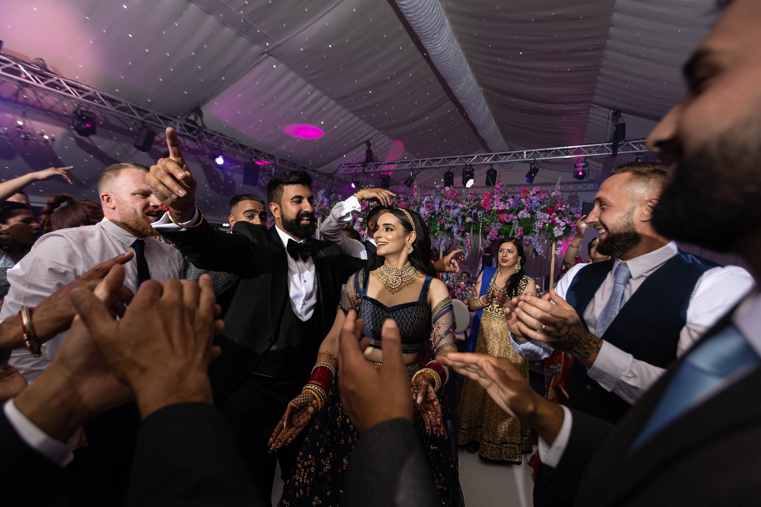 People dancing and celebrating at a wedding reception, with a bride in traditional Indian attire and guests dressed in formal clothing, under colorful stage lighting and floral decorations.