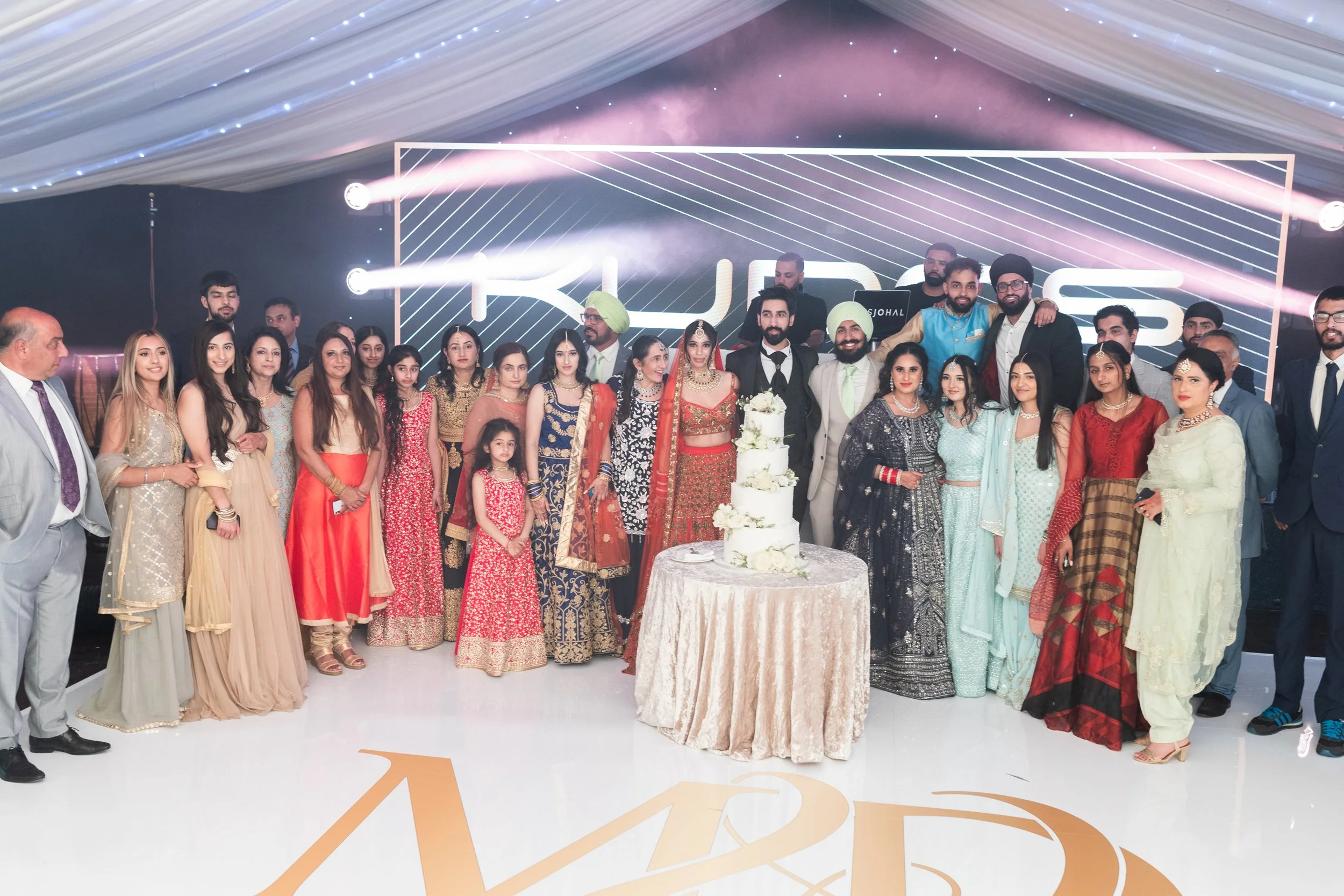 A large group of people dressed in traditional and formal attire gathered around a round table with a tall, tiered white wedding cake on it. The background features a large digital screen with bright lights and the text 'KUSH.'