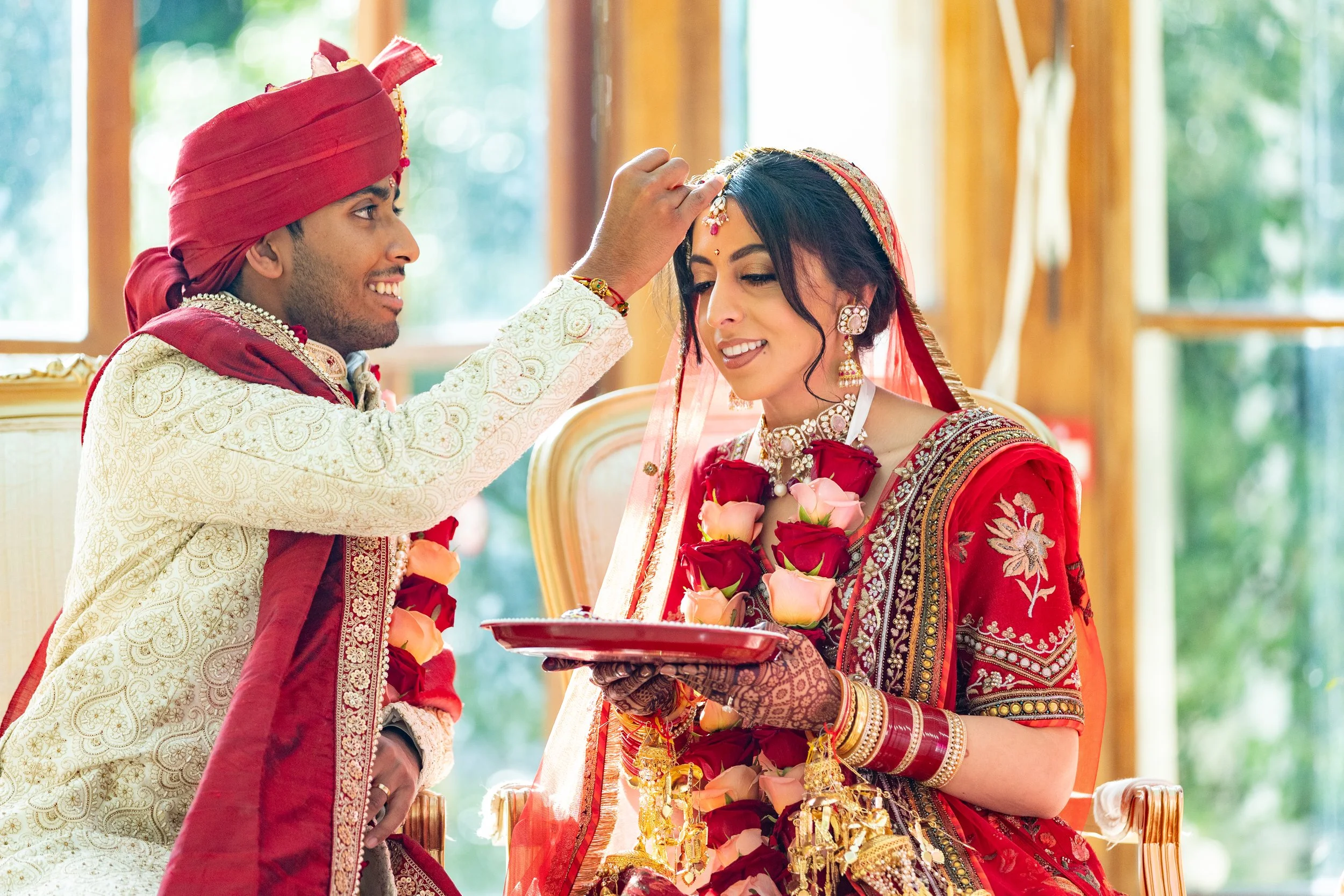 Indian bride and groom during wedding ceremony, with the groom placing a blessing on the bride's forehead, both dressed in traditional red and white attire, with floral jewelry and mehndi design on the bride's hands.