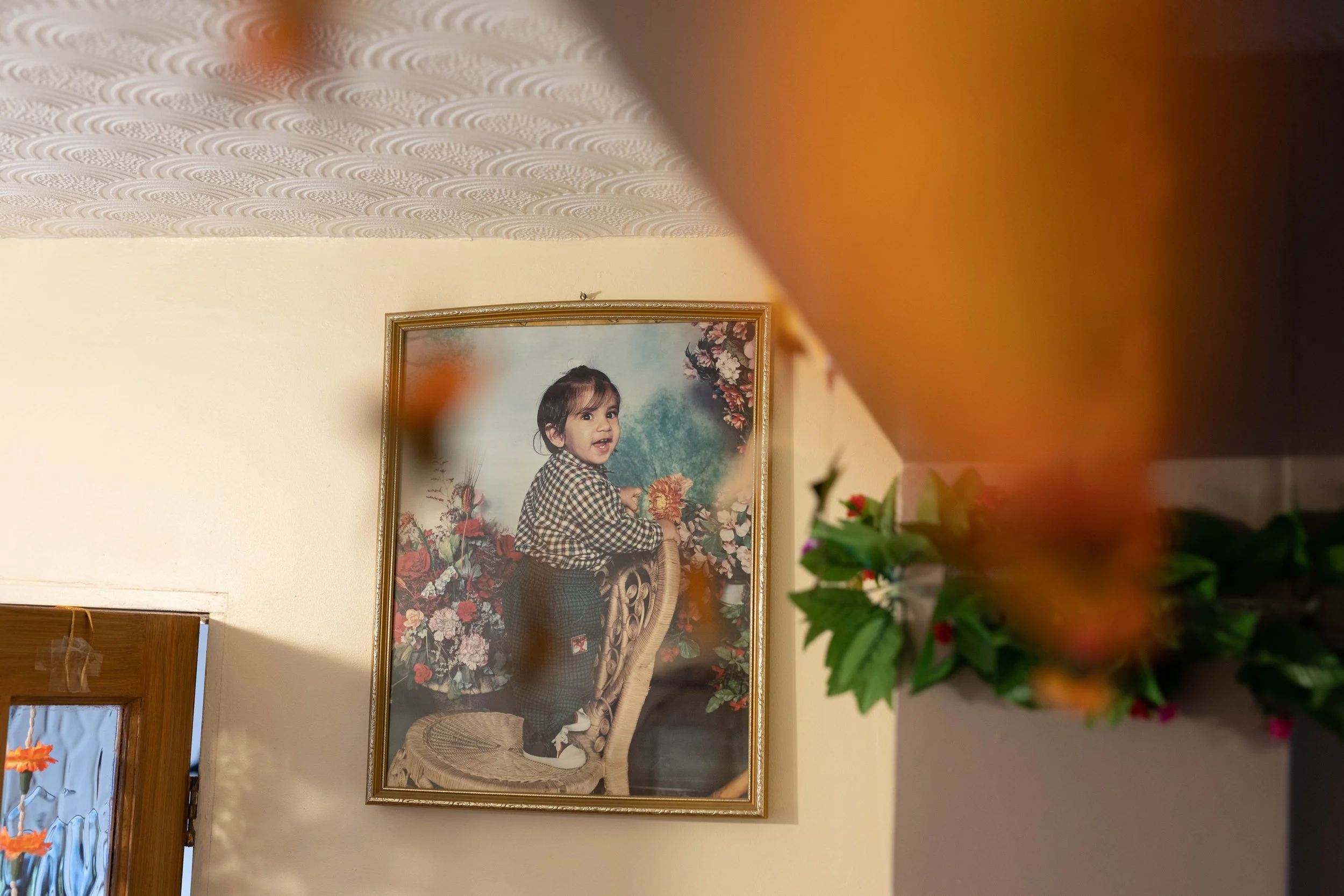 A framed photograph of a young girl standing on a chair, surrounded by floral decorations, hanging on a wall in a room with patterned ceiling and partially visible door with decorative glass.