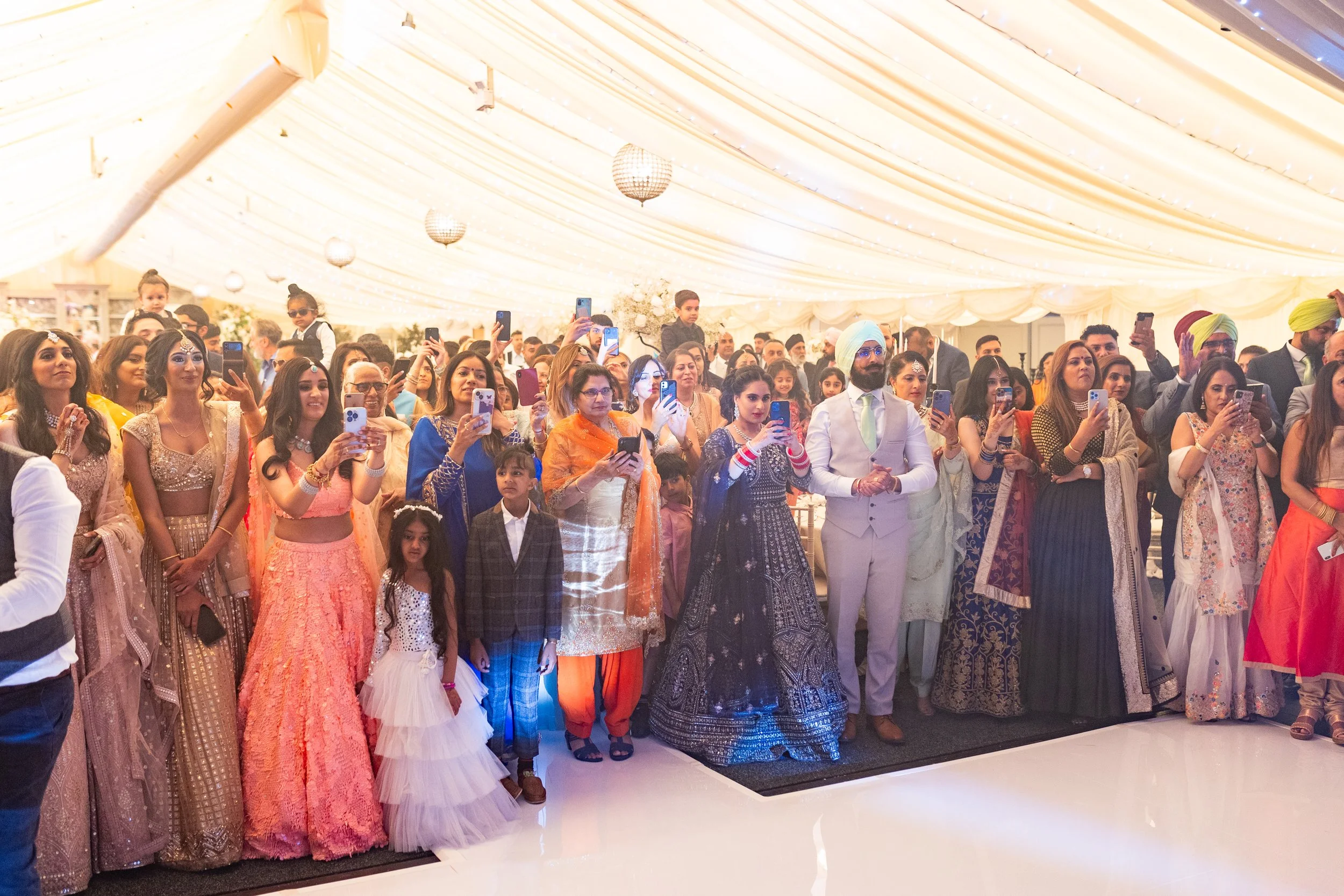 Group of people at a wedding reception under a decorated tent, many holding up phones to take photos. Men in traditional Indian attire, women in colorful sarees and dresses, children present, with a bright and festive atmosphere.