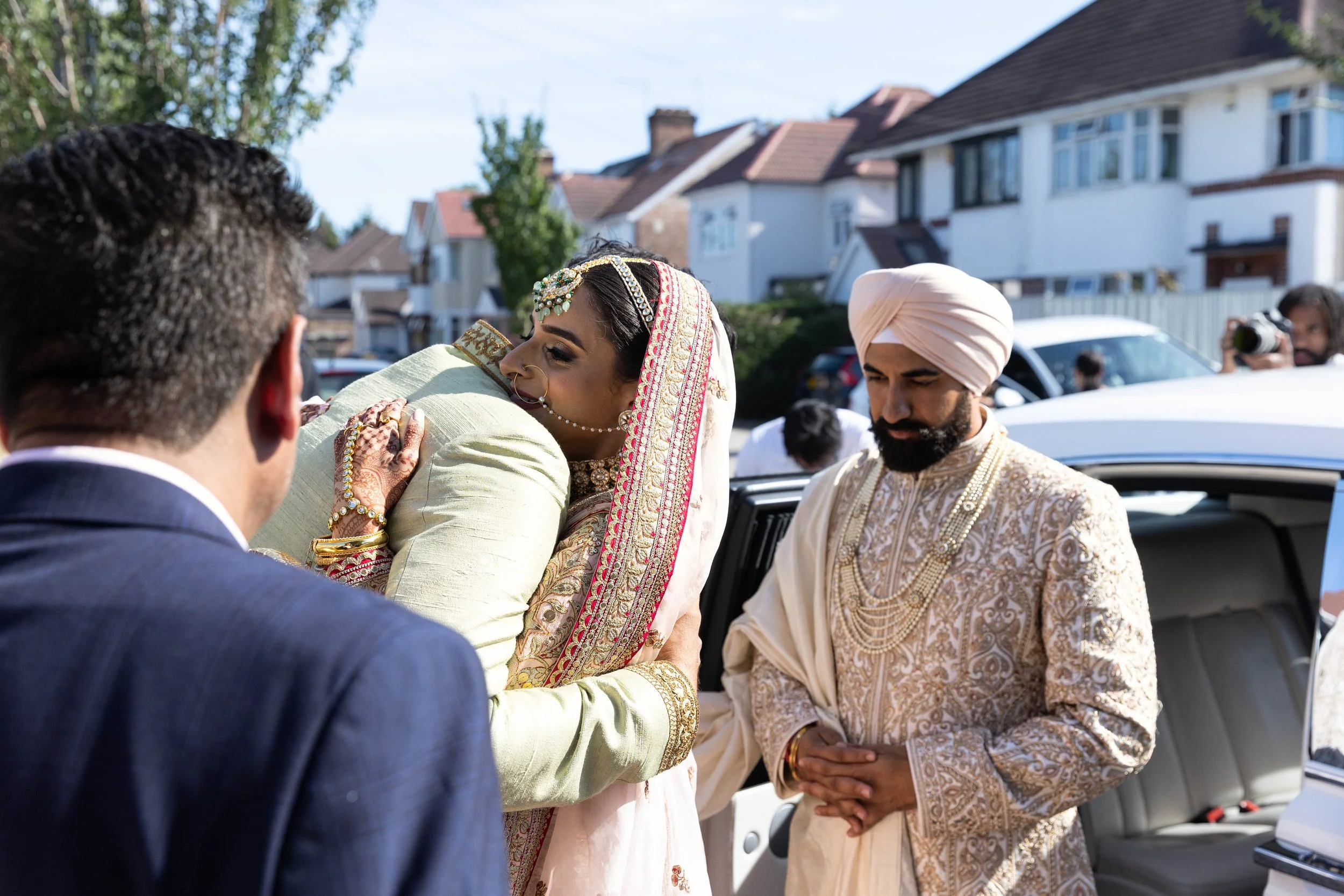 A bride in traditional Indian wedding attire hugging a guest, with a groom and other guests nearby, during a wedding celebration outside in a suburban neighborhood.