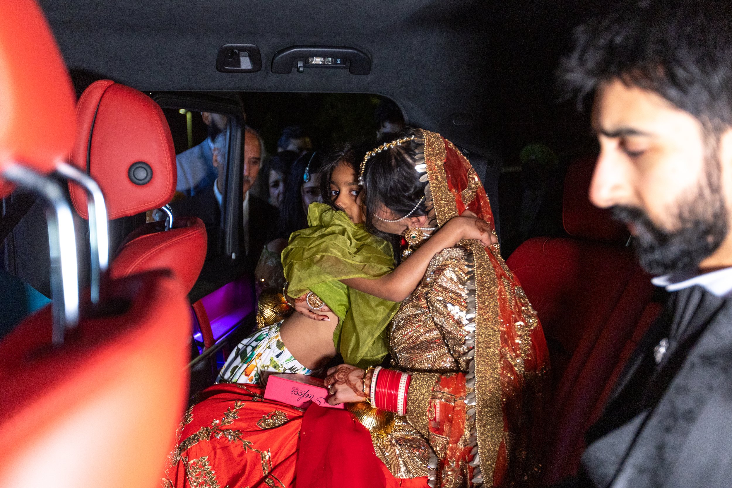 A woman dressed in traditional Indian wedding attire sharing a kiss with a young girl in a green outfit inside a car, with other people visible in the background.
