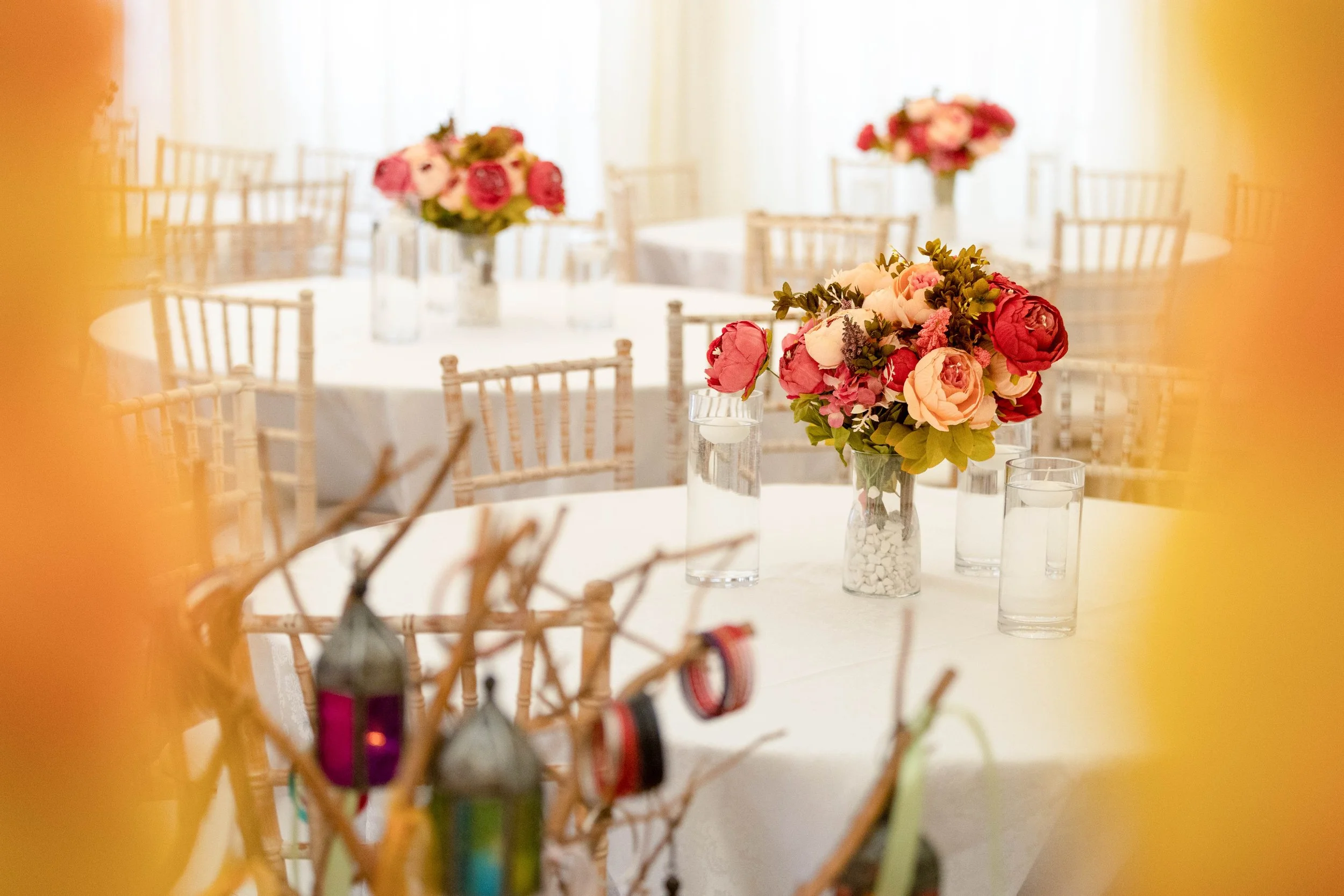 Elegant event space with round tables decorated with floral centerpieces featuring pink and red flowers in glass vases. Clear glasses of water are on the tables, surrounded by beige chiavari chairs. Bright natural light filters through sheer curtains