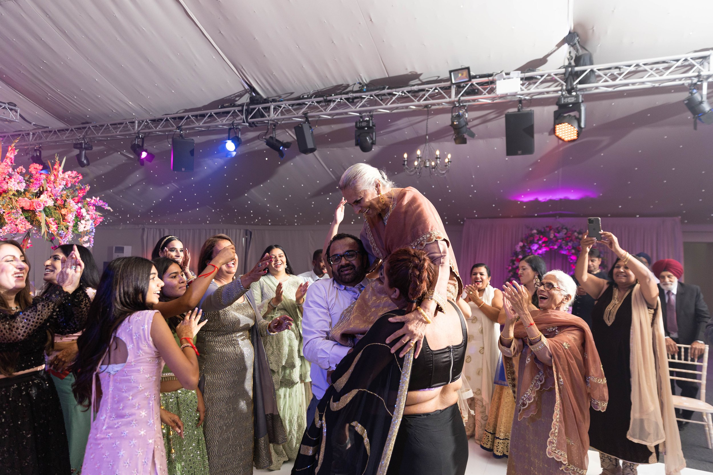 Elderly woman being lifted by a man at a celebration with friends and family, diverse group of people dressed in traditional and festive clothing, colorful floral arrangements, and decorative lighting.
