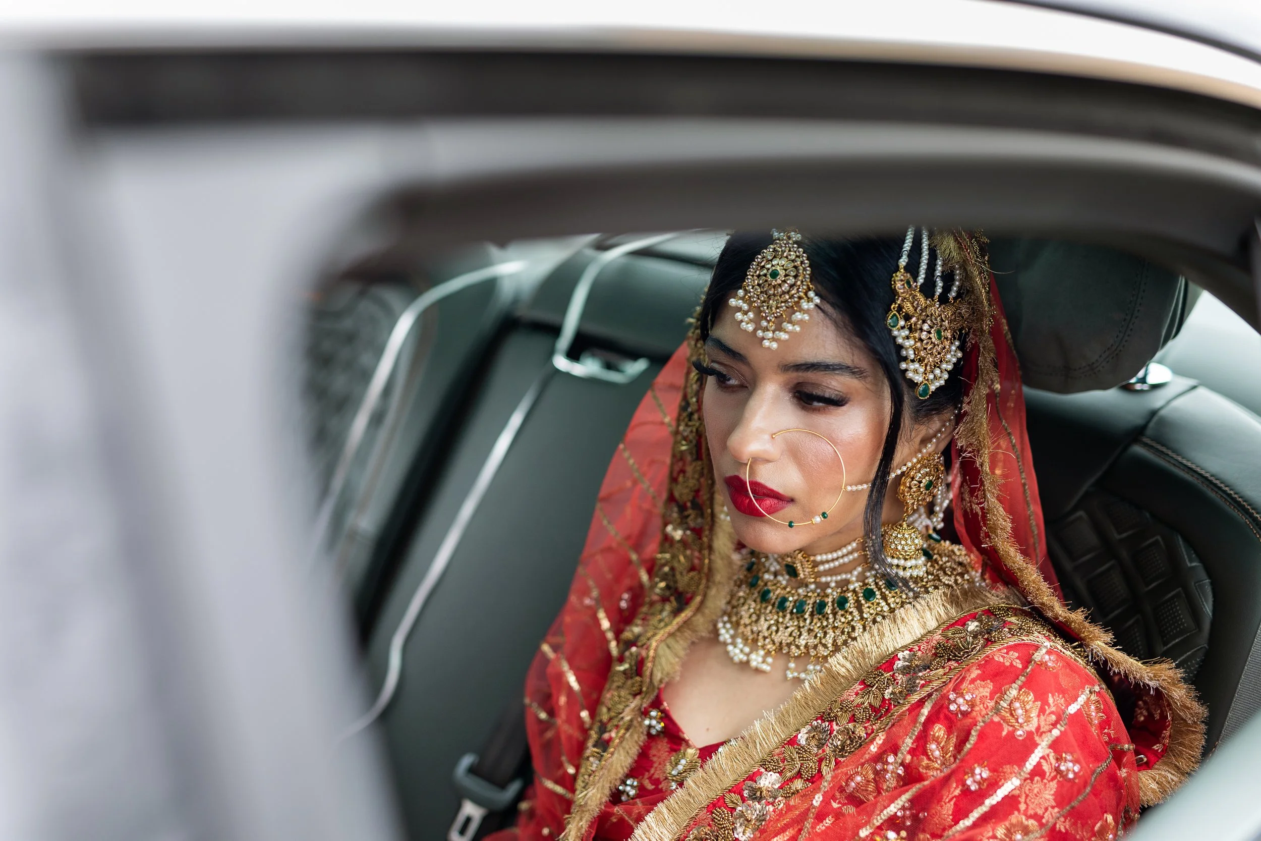 A woman dressed in traditional Indian wedding attire, wearing red and gold jewelry, including a necklace, earrings, and headpiece, sitting in the backseat of a car.