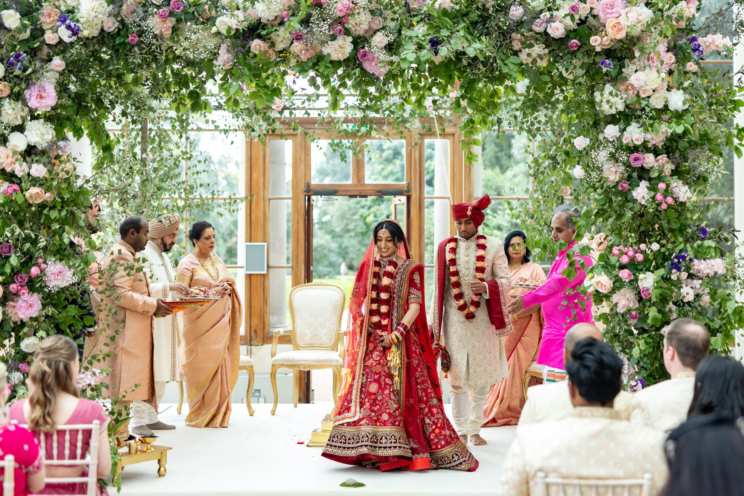 An Indian wedding ceremony with a bride and groom dressed in traditional red and white attire, standing under a floral arch, surrounded by family members, with guests seated and participating in the celebration.