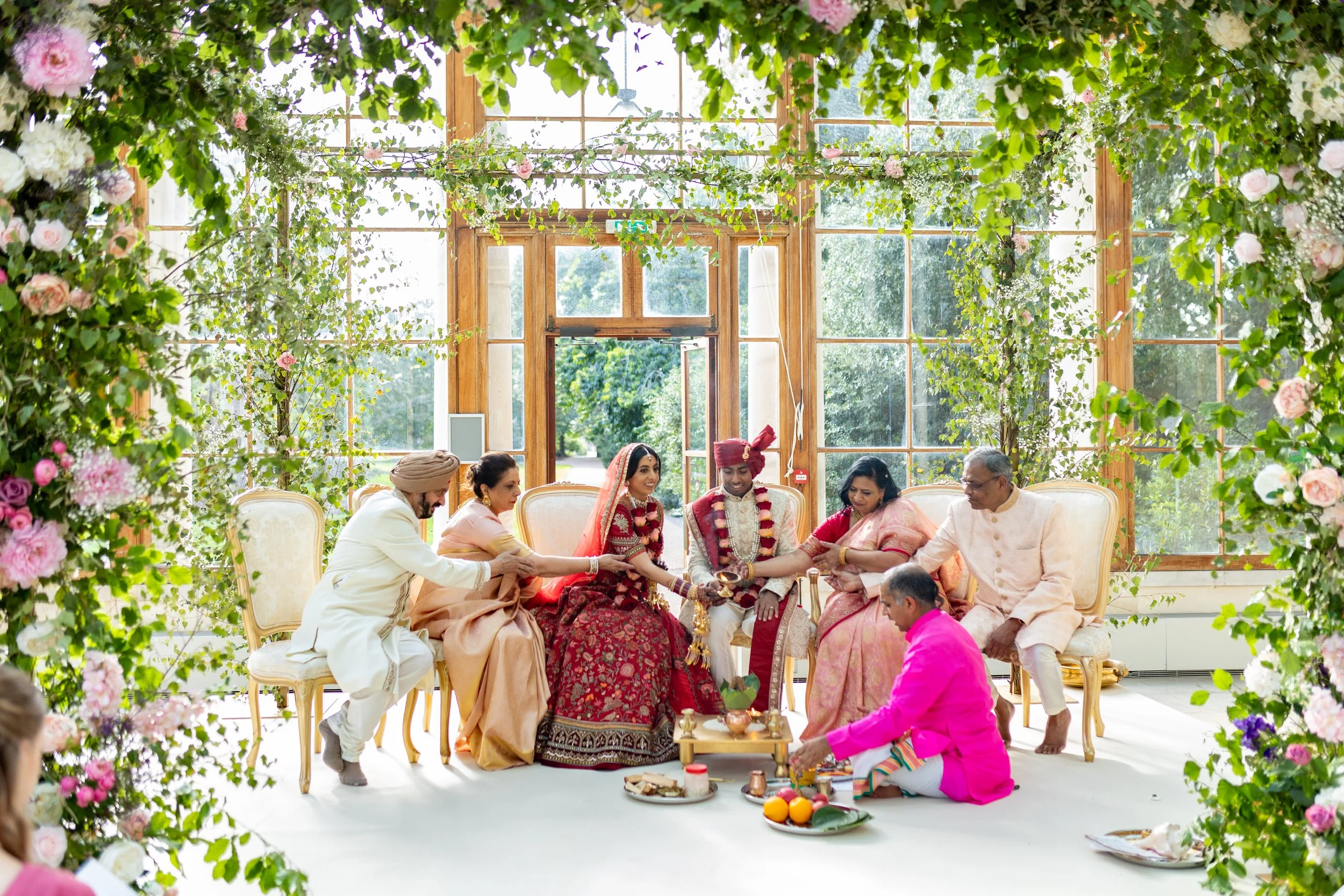 Indian wedding ceremony with couple and family members seated inside a bright, floral-decorated hall, participating in a traditional ritual.